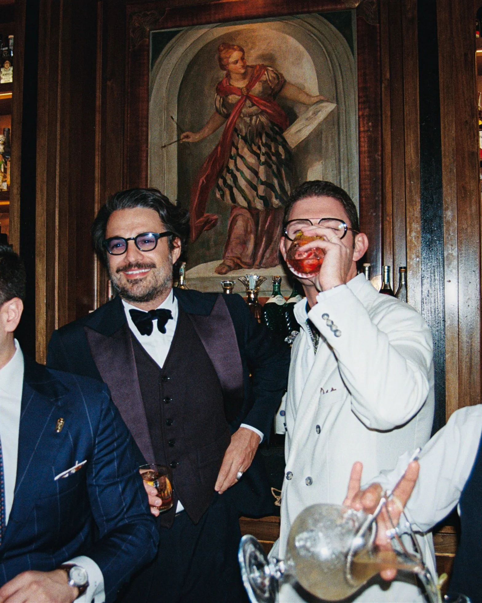Two men in formal attire at a bar, one holding a drink and the other drinking from a glass, with a painting of a woman holding a staff in the background.