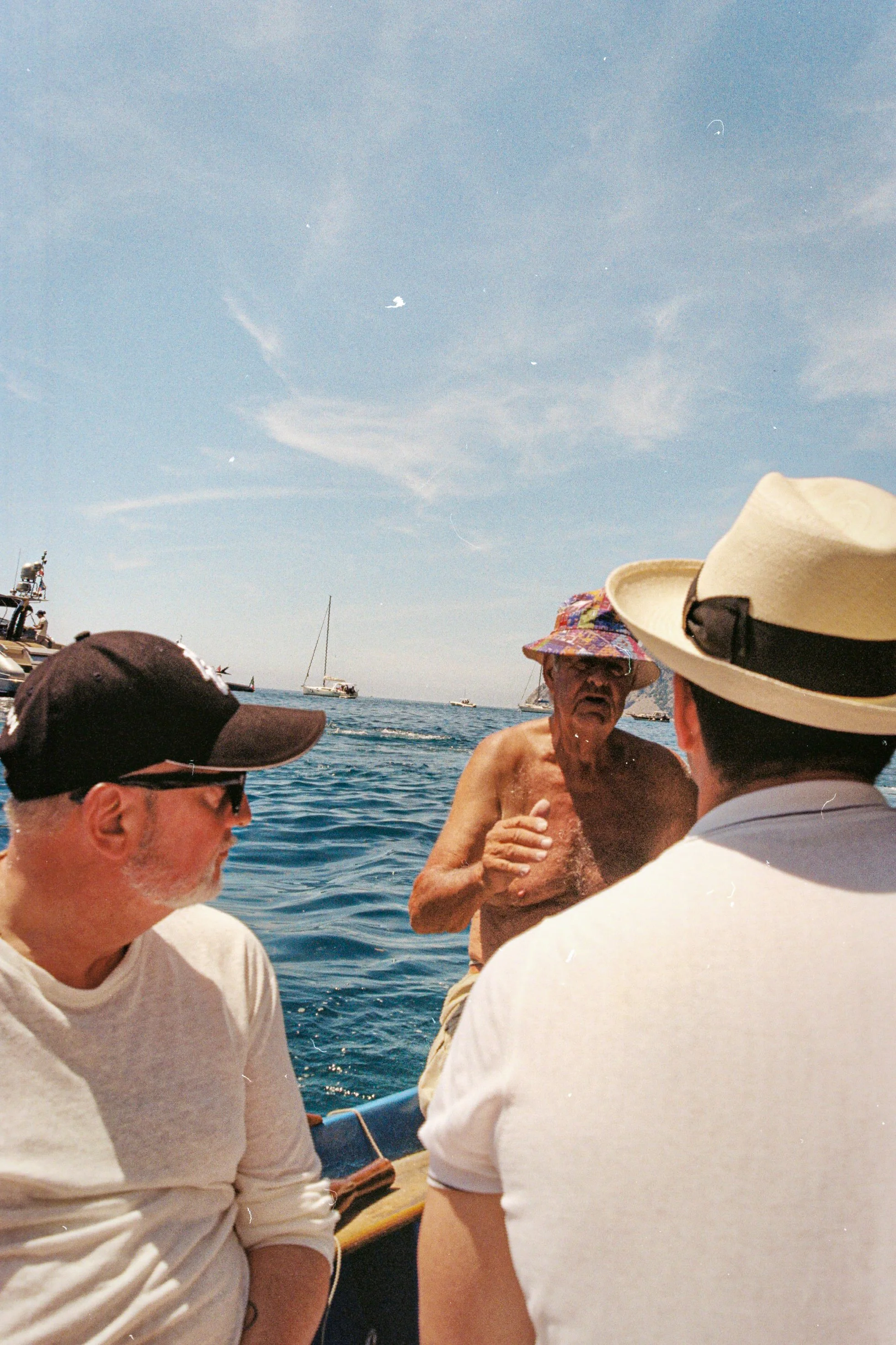Three men on a boat, one shirtless wearing a colorful hat, talking to two others wearing hats and sunglasses, with the ocean and sailboats in the background under a partly cloudy sky.