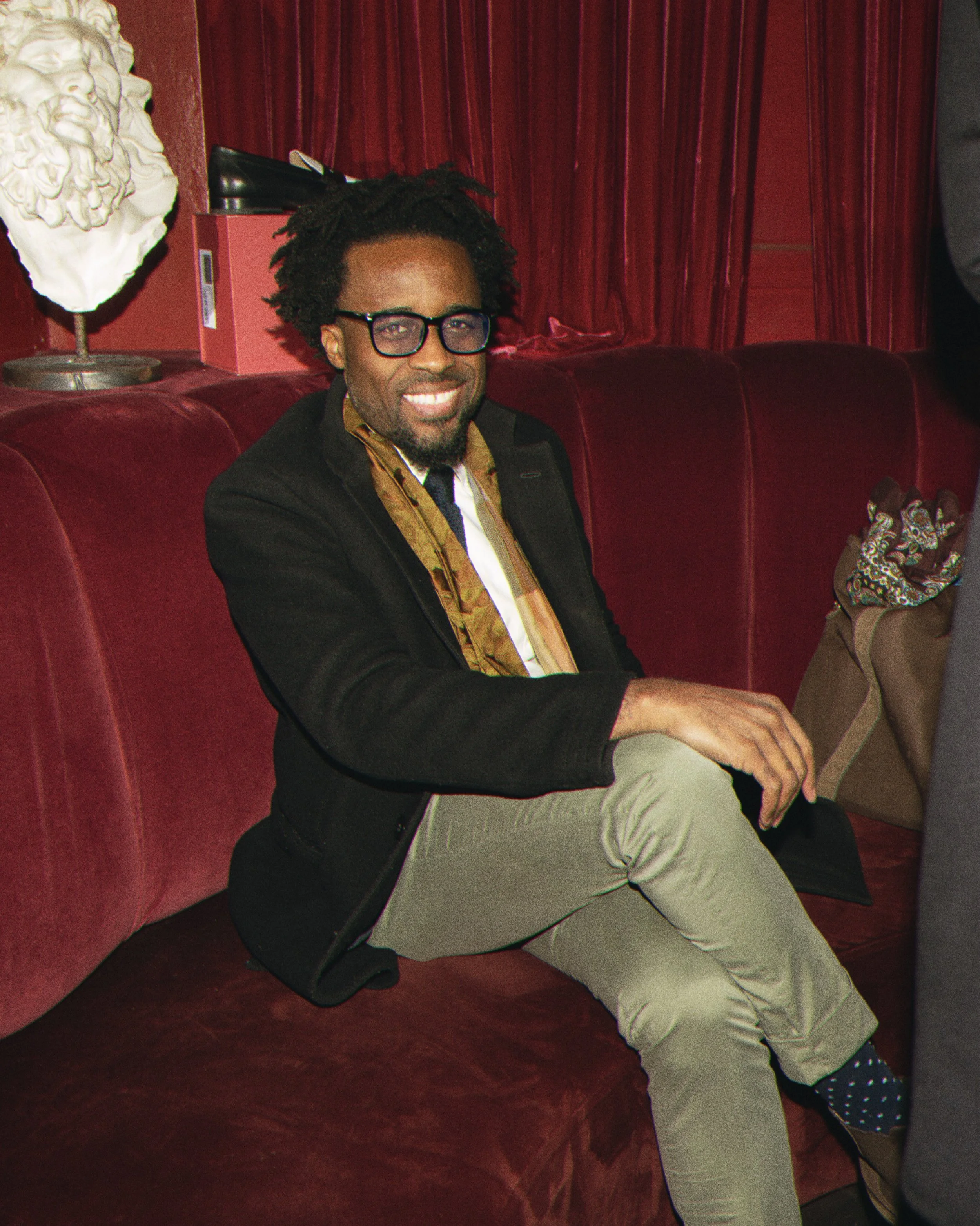 A man with curly hair, glasses, and a beard sitting on a red velvet couch, smiling at the camera. He is wearing a black blazer, a patterned shirt, and beige pants. There is a white bust sculpture on a stand and a pink box with some objects on it in t