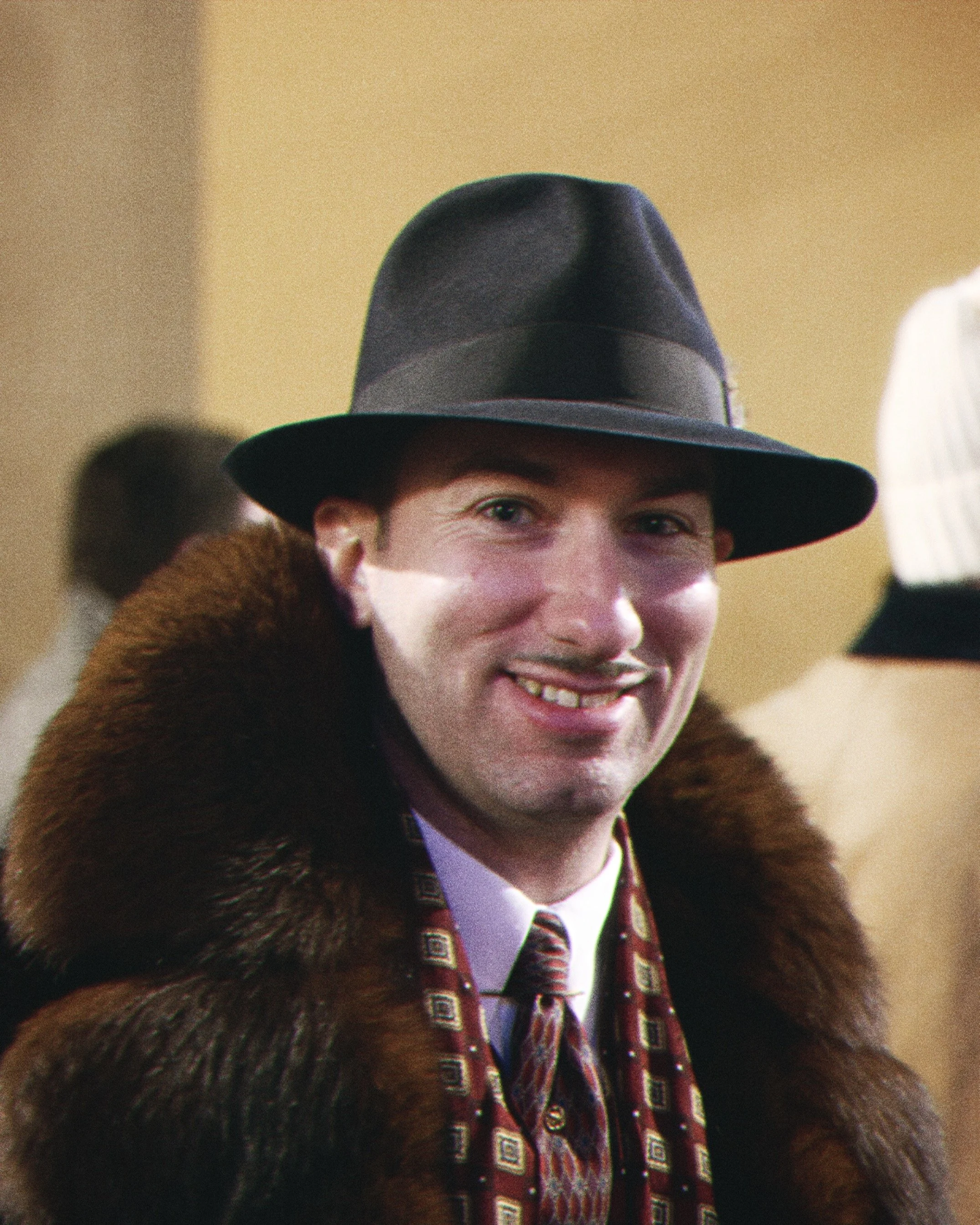 A person smiling, wearing a dark fedora hat and a patterned red and black tie, with a fur coat over a white shirt.