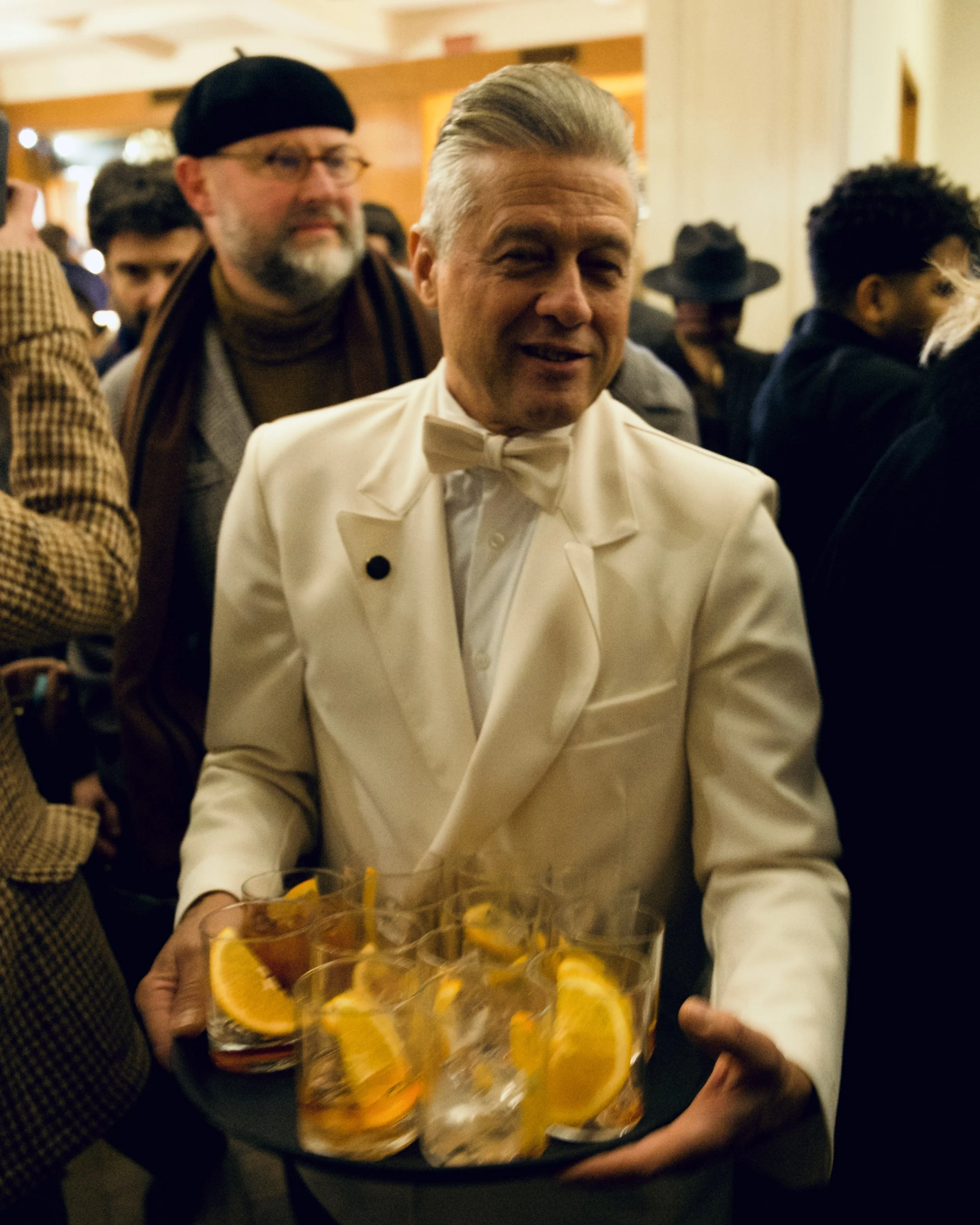 A man in a white tuxedo with a bow tie holding a tray of orange cocktails garnished with orange slices at a social gathering or party.