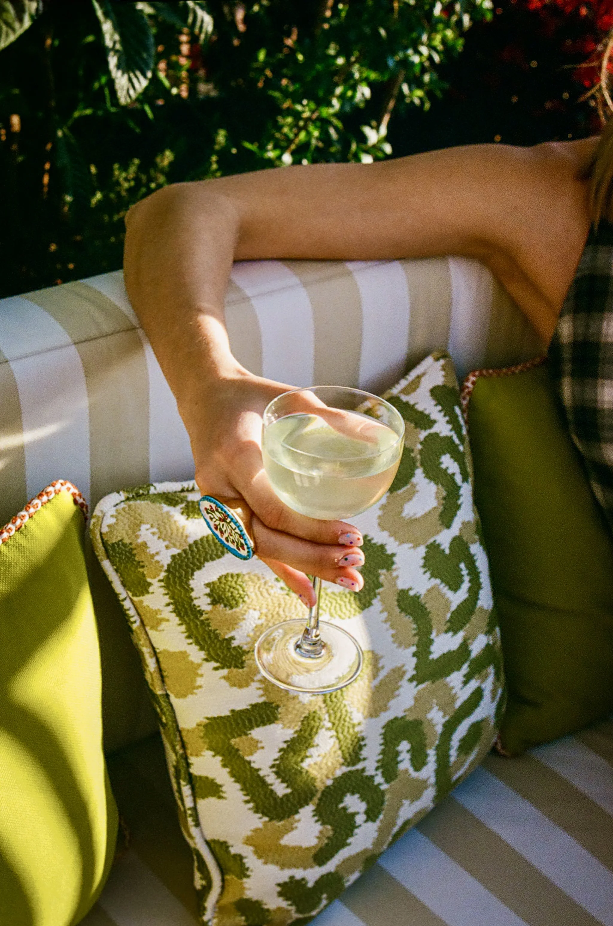 Person holding a glass of white wine sitting outdoors on a patterned fabric sofa with green foliage in the background.
