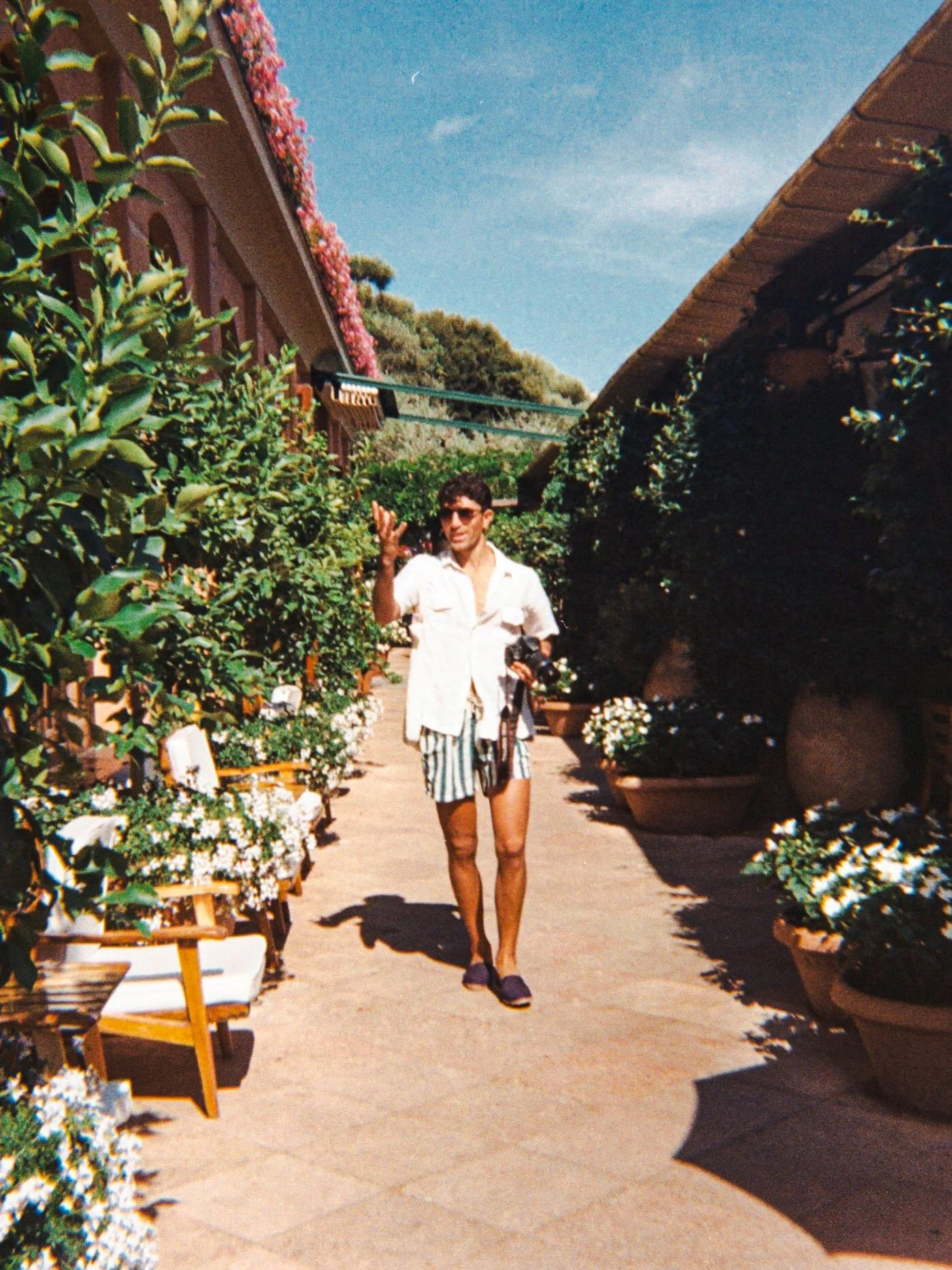 A man in a white shirt and striped shorts walking through a garden pathway with planters of flowers on both sides. The sky is clear with a visible moon and a small cloud. The area is shaded by large plants and there are chairs along the path.