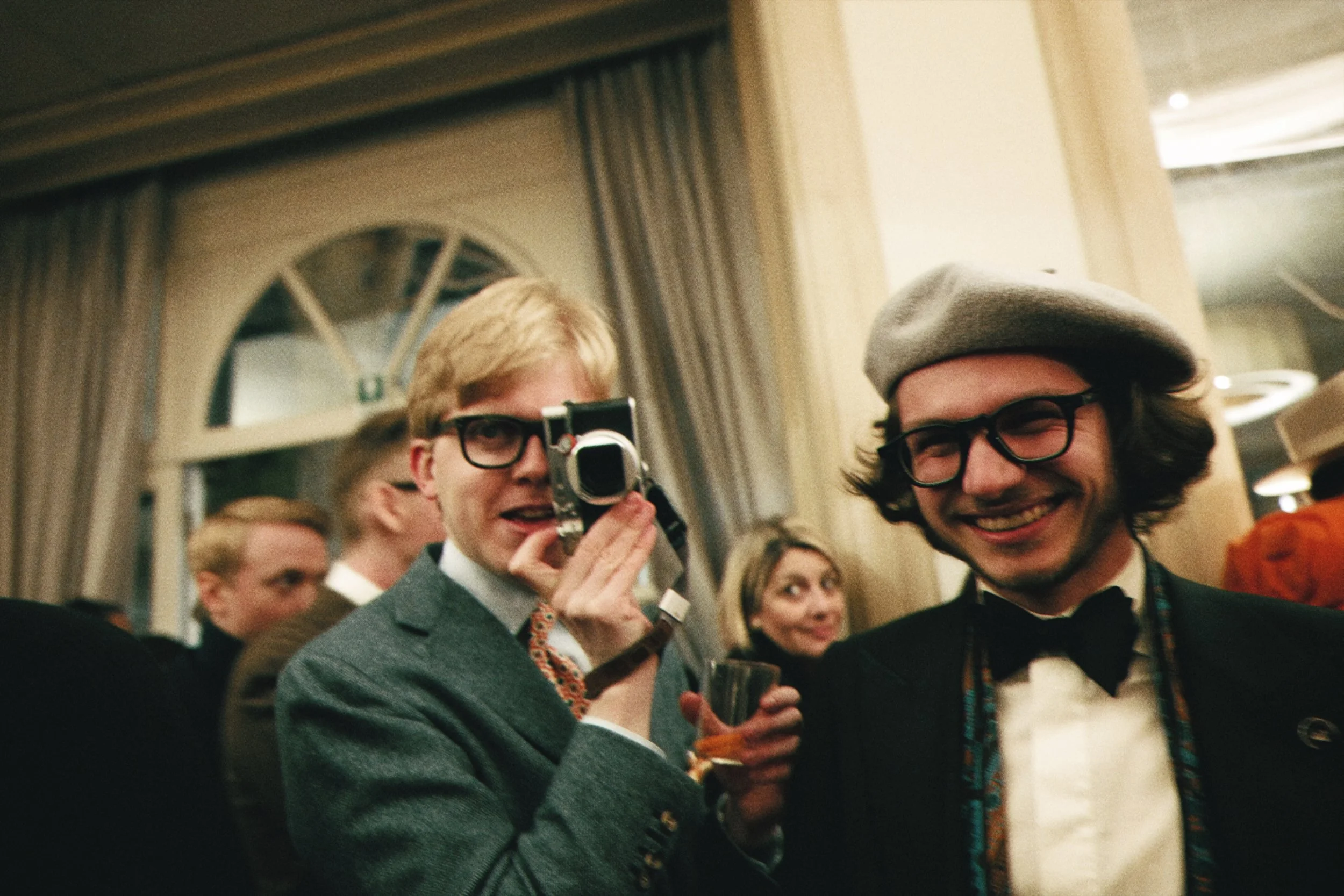 Two men in formal attire smiling at a social gathering, one holding a camera, in an elegant indoor setting with high ceilings and decorative curtains.