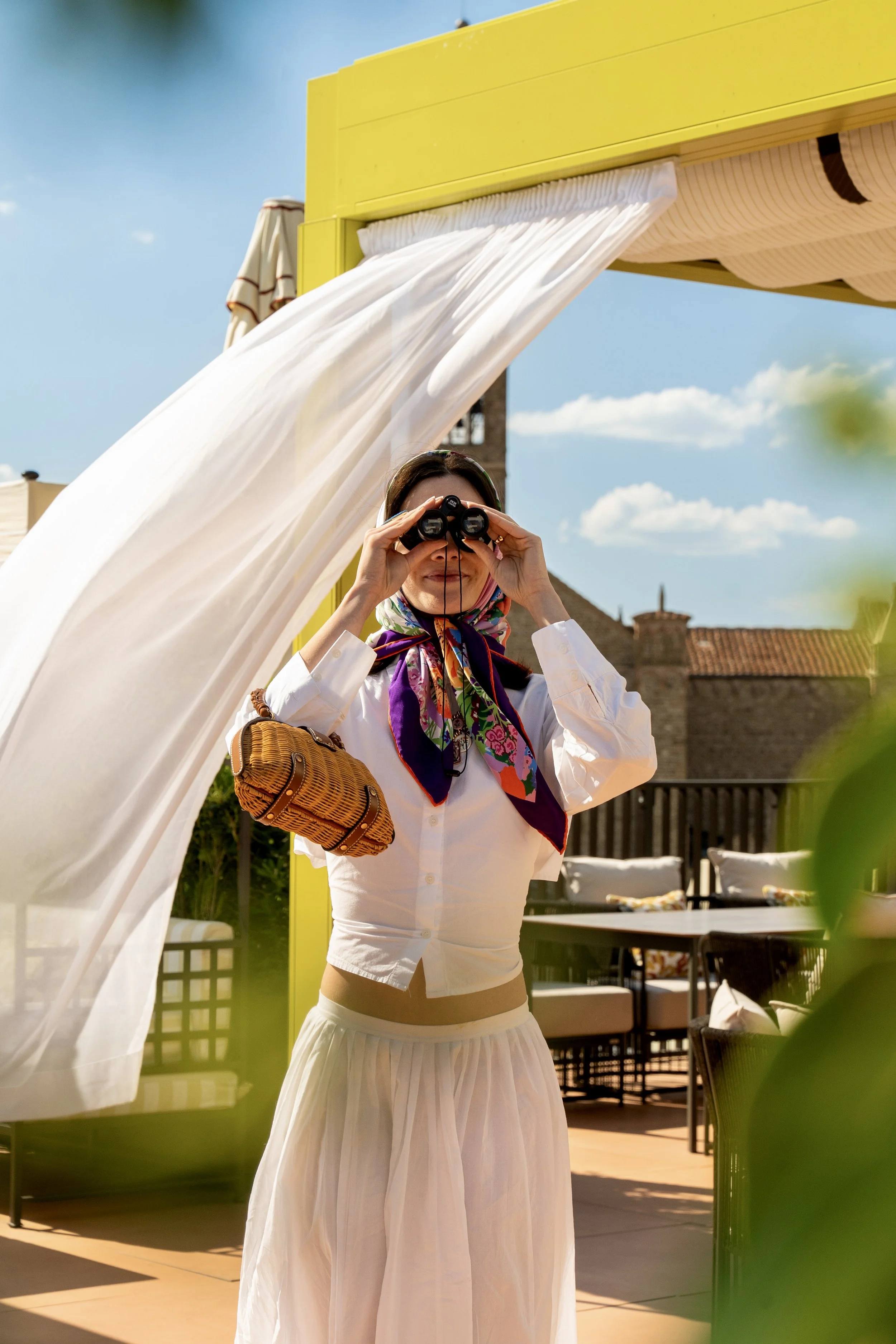 A woman is standing outdoors on a sunny day, looking through binoculars. She is wearing a white blouse, a patterned scarf, and a white skirt. There is a yellow structure with white curtains flowing in the breeze behind her, along with outdoor furnitu