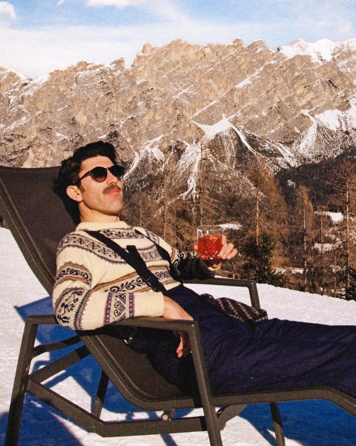 A man with a mustache and sunglasses lounging on a chair outdoors in a snowy mountainous landscape, holding a glass of red beverage.