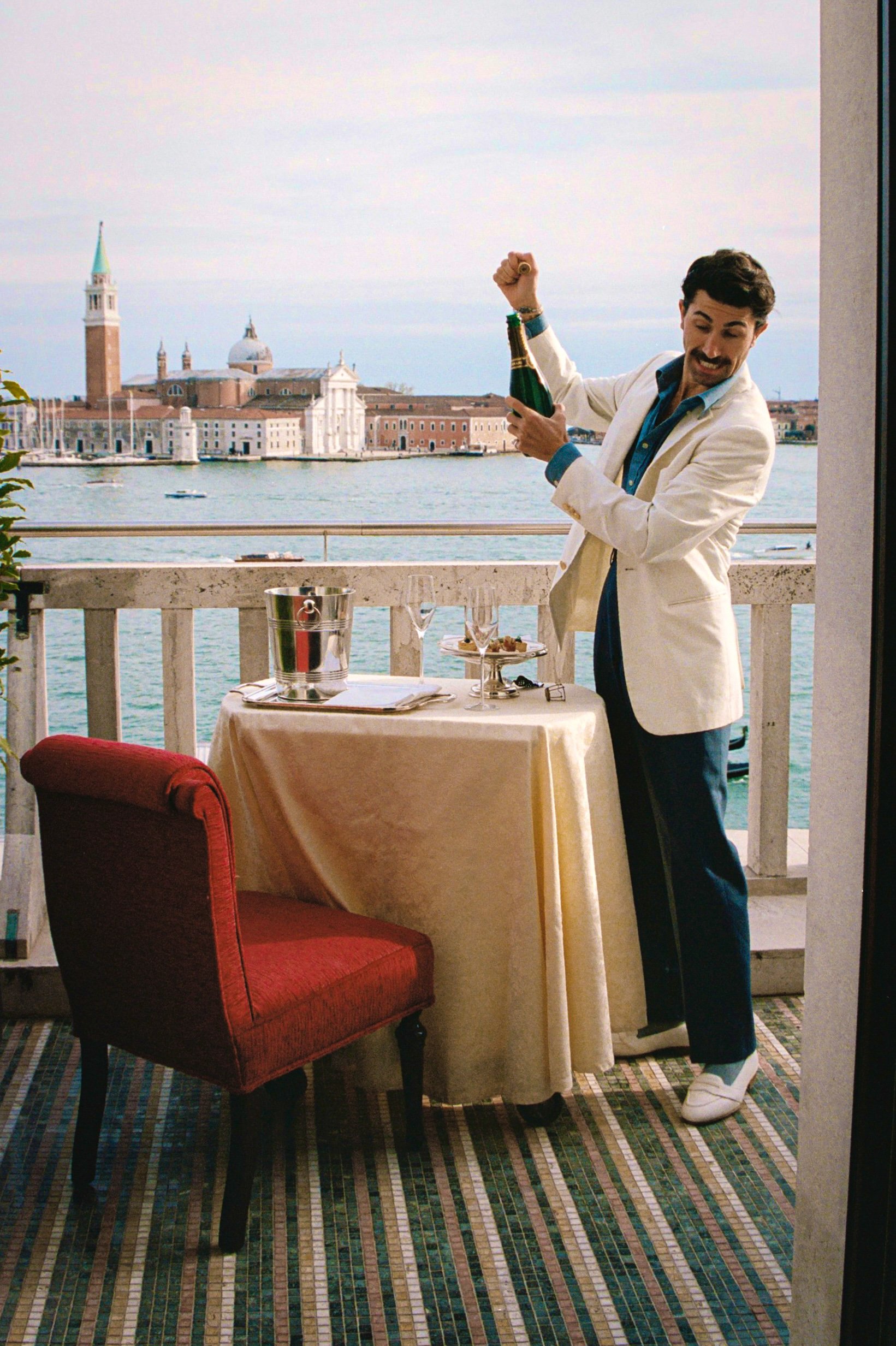 A man in a white blazer and dark pants opening a bottle of champagne on a balcony overlooking a waterway and historic buildings in Venice, Italy.