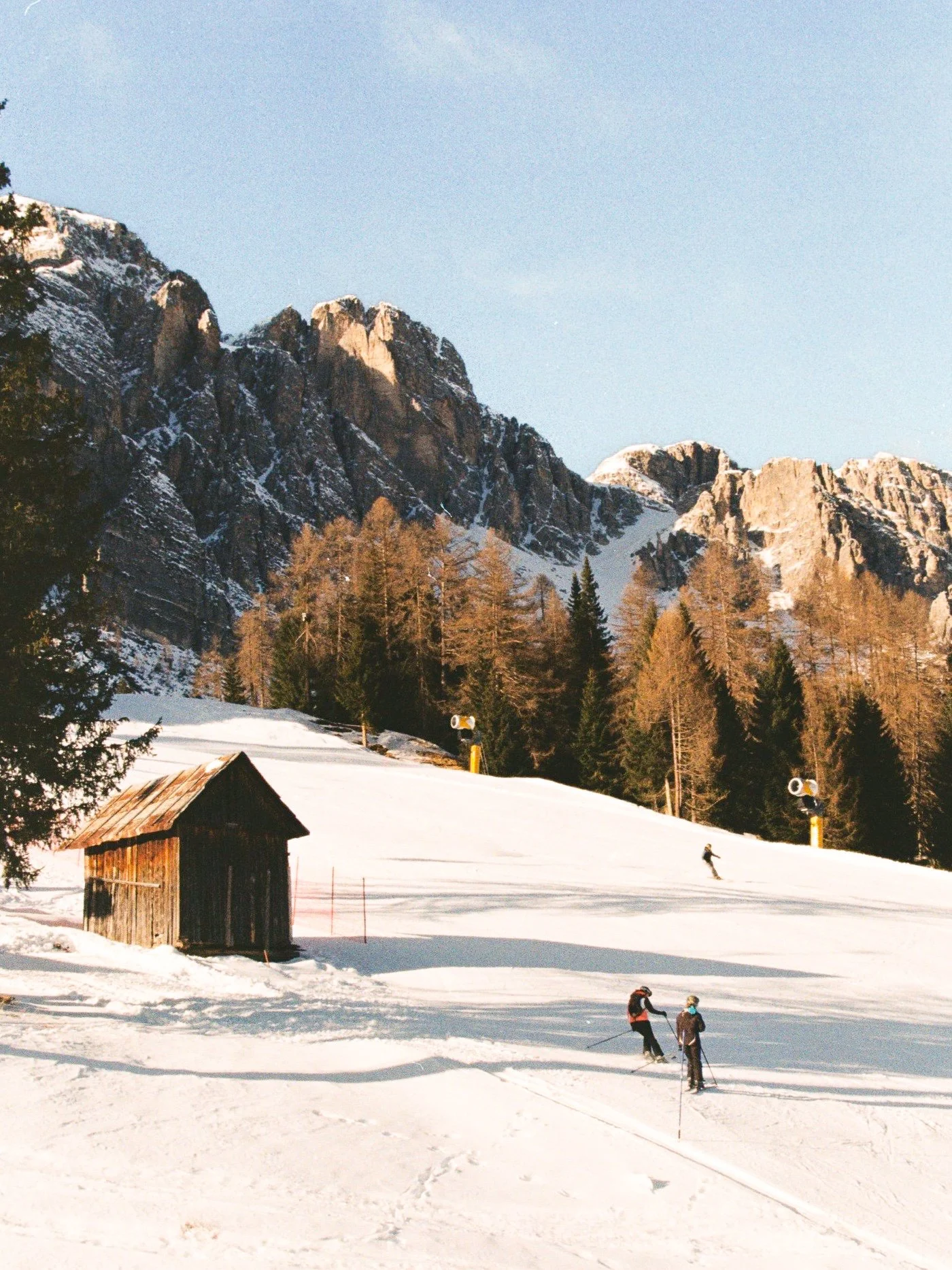 Skiers on a snowy mountain slope with wooden cabin, trees, mountains, and clear sky in the background.