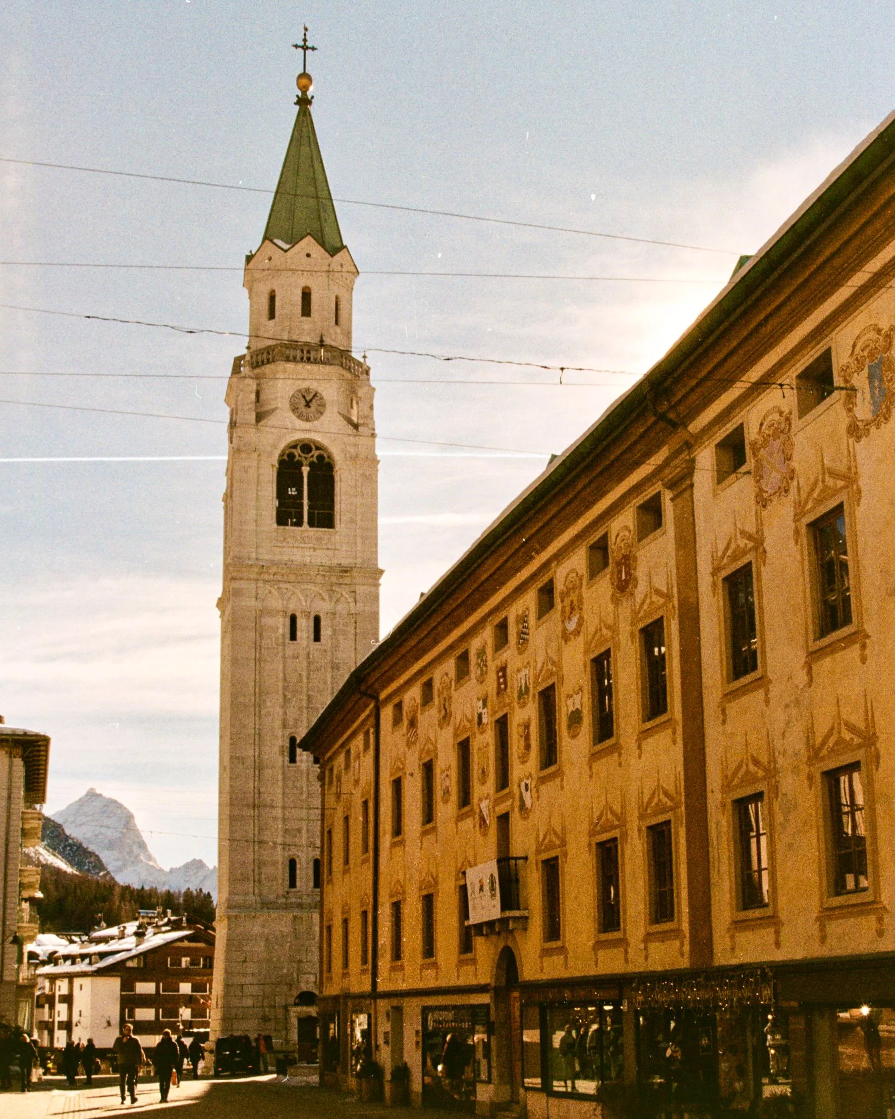 Tall clock tower with a green spire and a cross on top, next to a yellow building with painted emblems on the wall, in a town square with people walking and snow-capped mountains in the background.