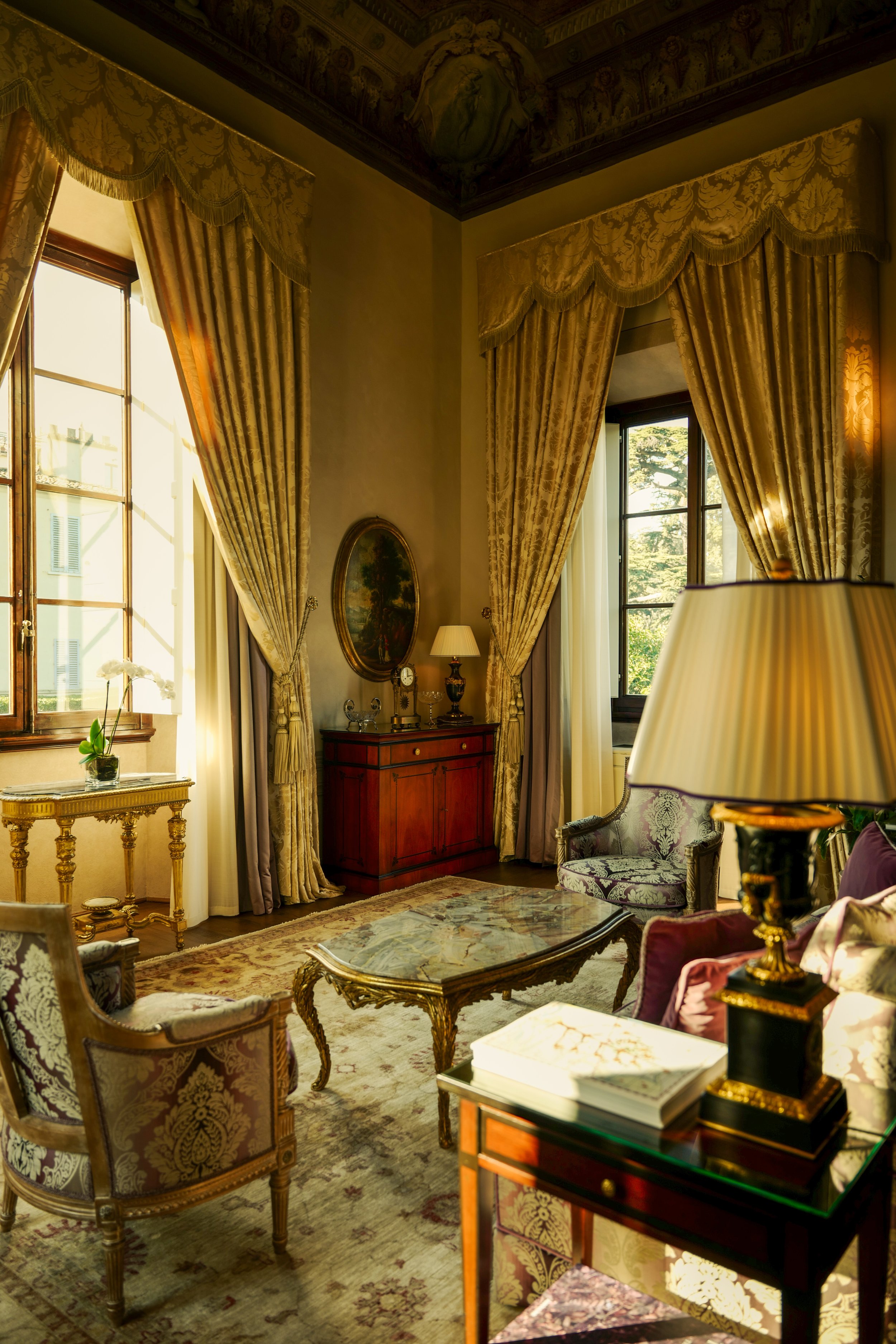 Elegant vintage living room with gold and floral upholstered furniture, ornate curtains, large windows, a marble-topped coffee table, and decorative lamps.