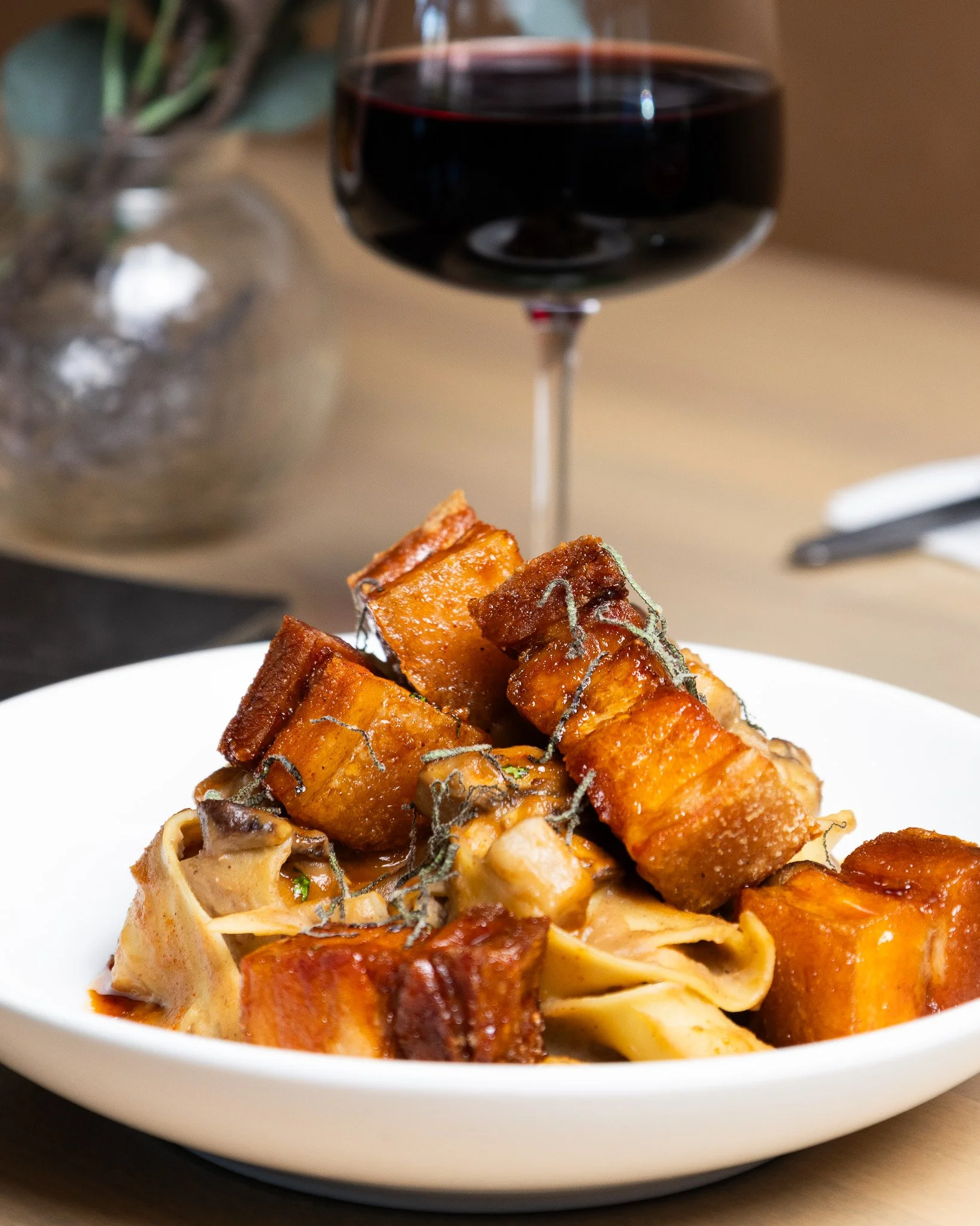 A white plate of baked pasta topped with pork belly, garnished with herbs, and accompanied by a glass of red wine in the background, with a blurred plant on the left side.