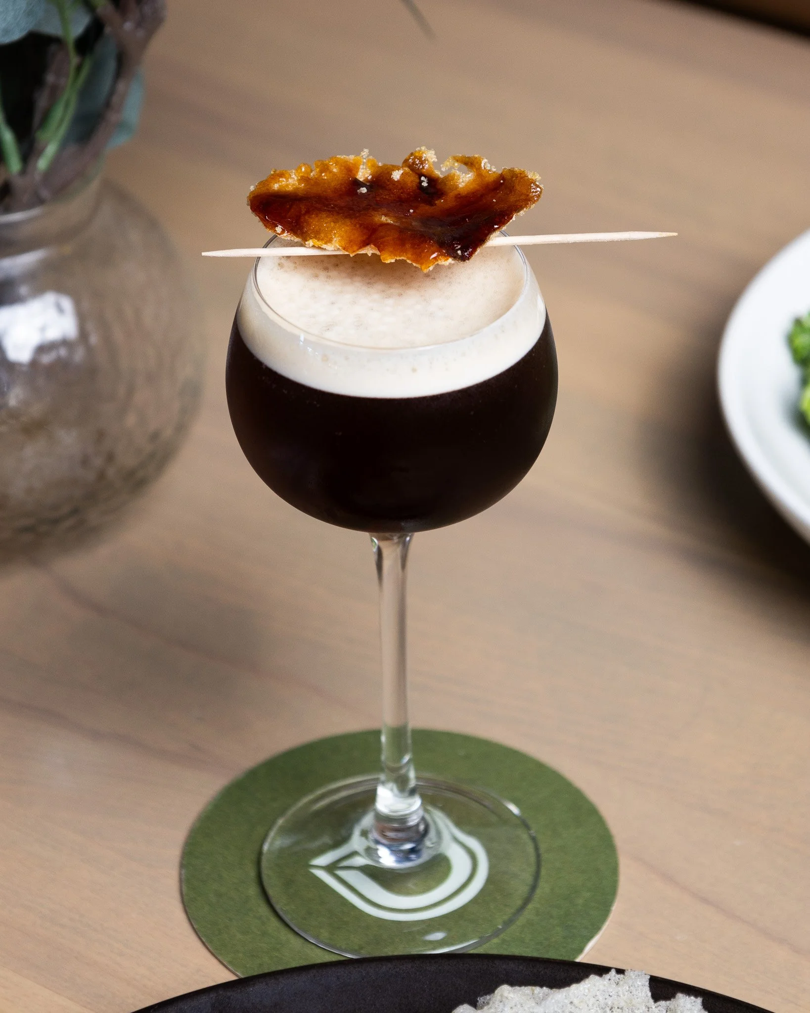 A dark cocktail, topped with a small piece of caramelized sugar on a toothpick, placed on a green coaster on a wooden table.