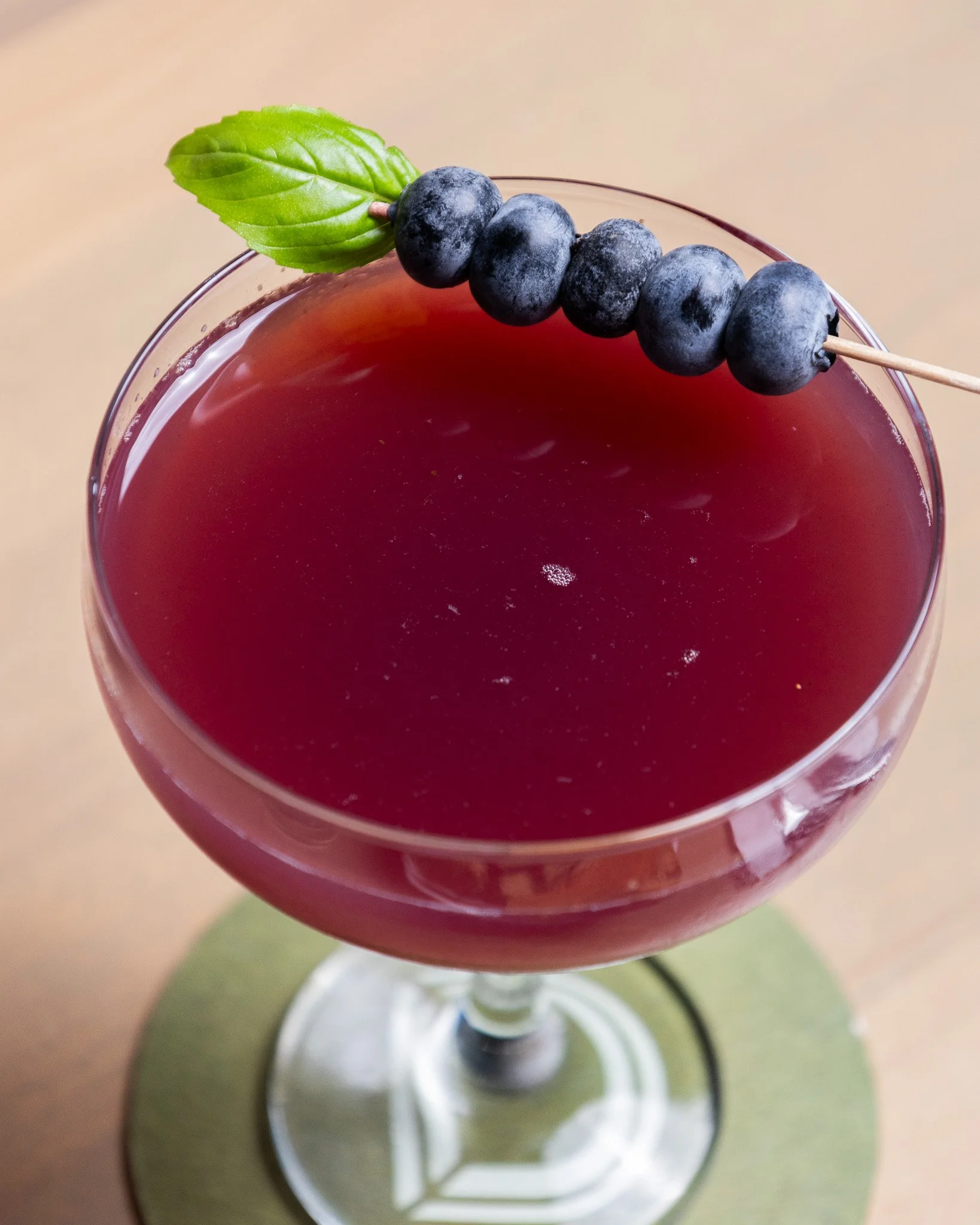 Close-up view of a red beverage in a glass with a skewer of blueberries and a green basil leaf garnish.