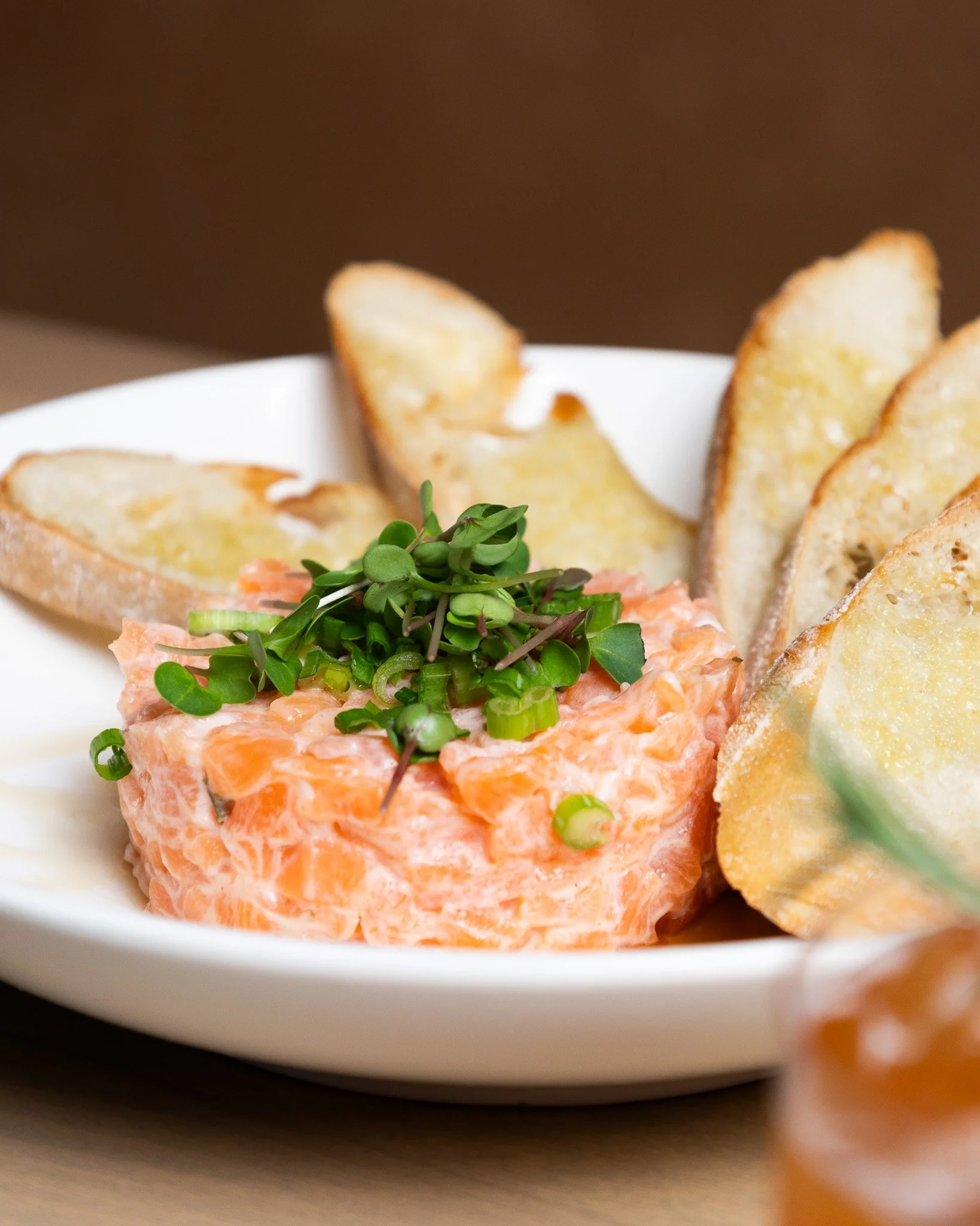 Close-up of a white bowl filled with salmon tartare topped with microgreens, with toasted bread slices in the background.