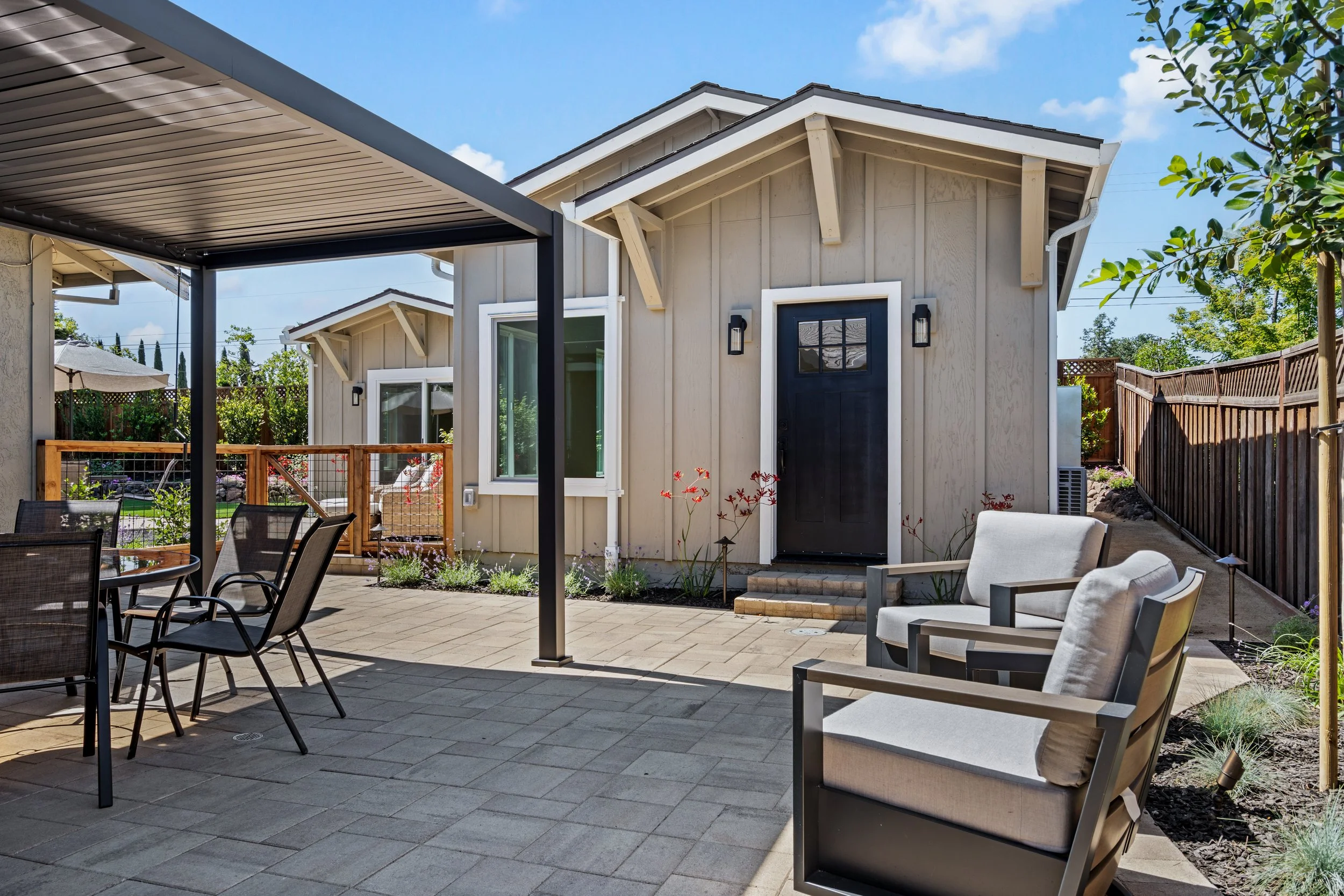backyard patio with outdoor seating area, including chairs and a dining table, in front of a small light-colored house with a dark front door and a fence around the yard.