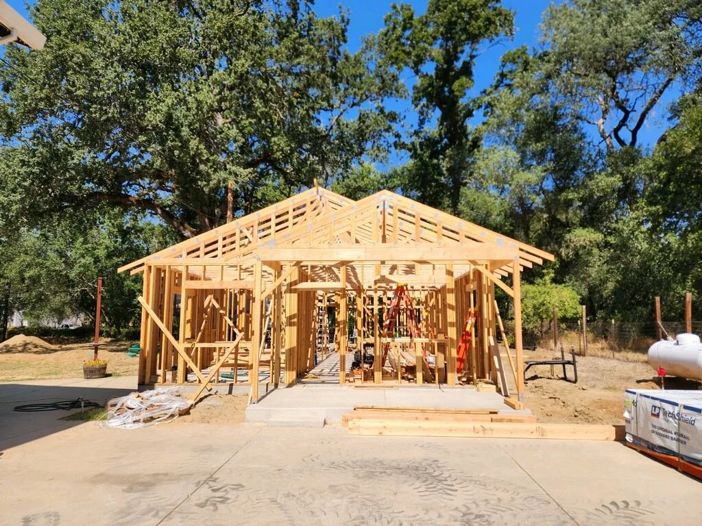 A house under construction with wooden framing, set against green trees and a clear blue sky.