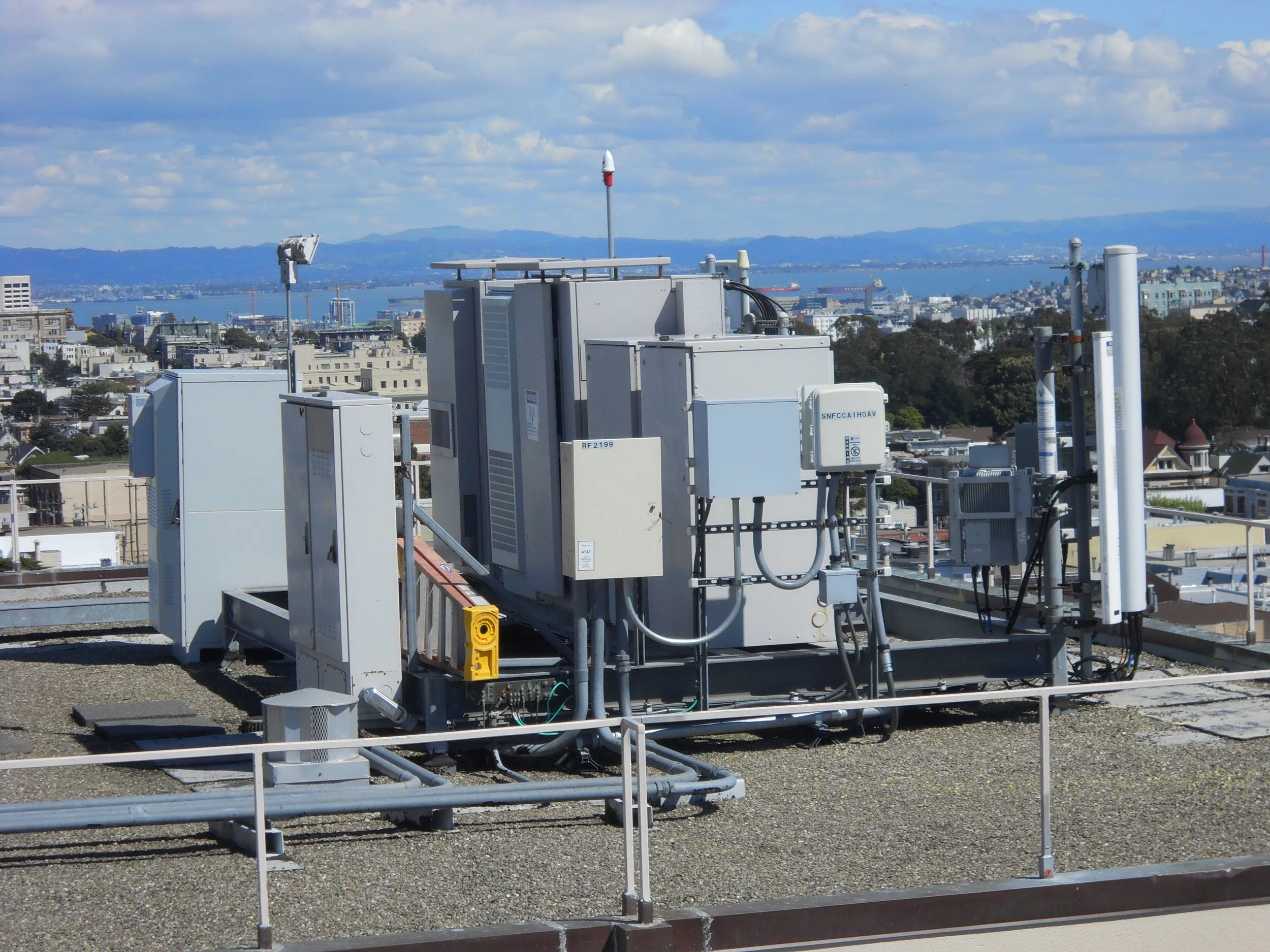 Rooftop view with various electrical and communication equipment, with cityscape and water in the background.