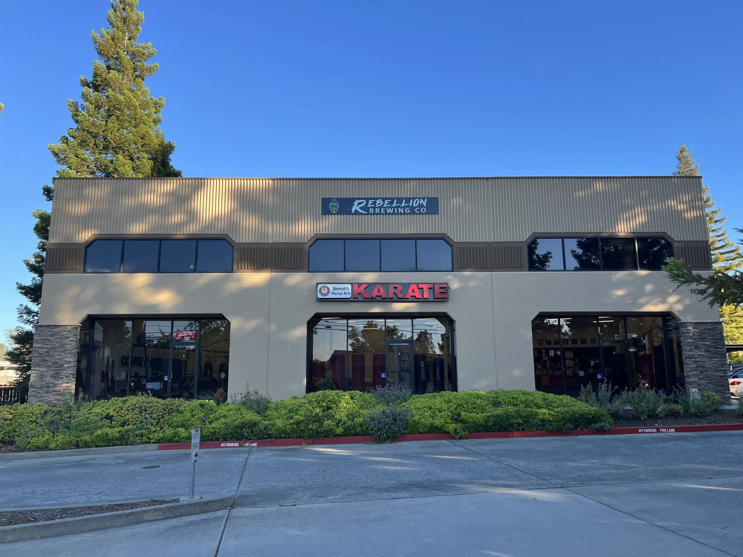 Front view of a two-story commercial building with a sign for Rebellion Brewing Co on the upper level and a martial arts dojo, Bennett's Martial Arts, on the lower level, with a landscaped flower bed and parking lot in front.