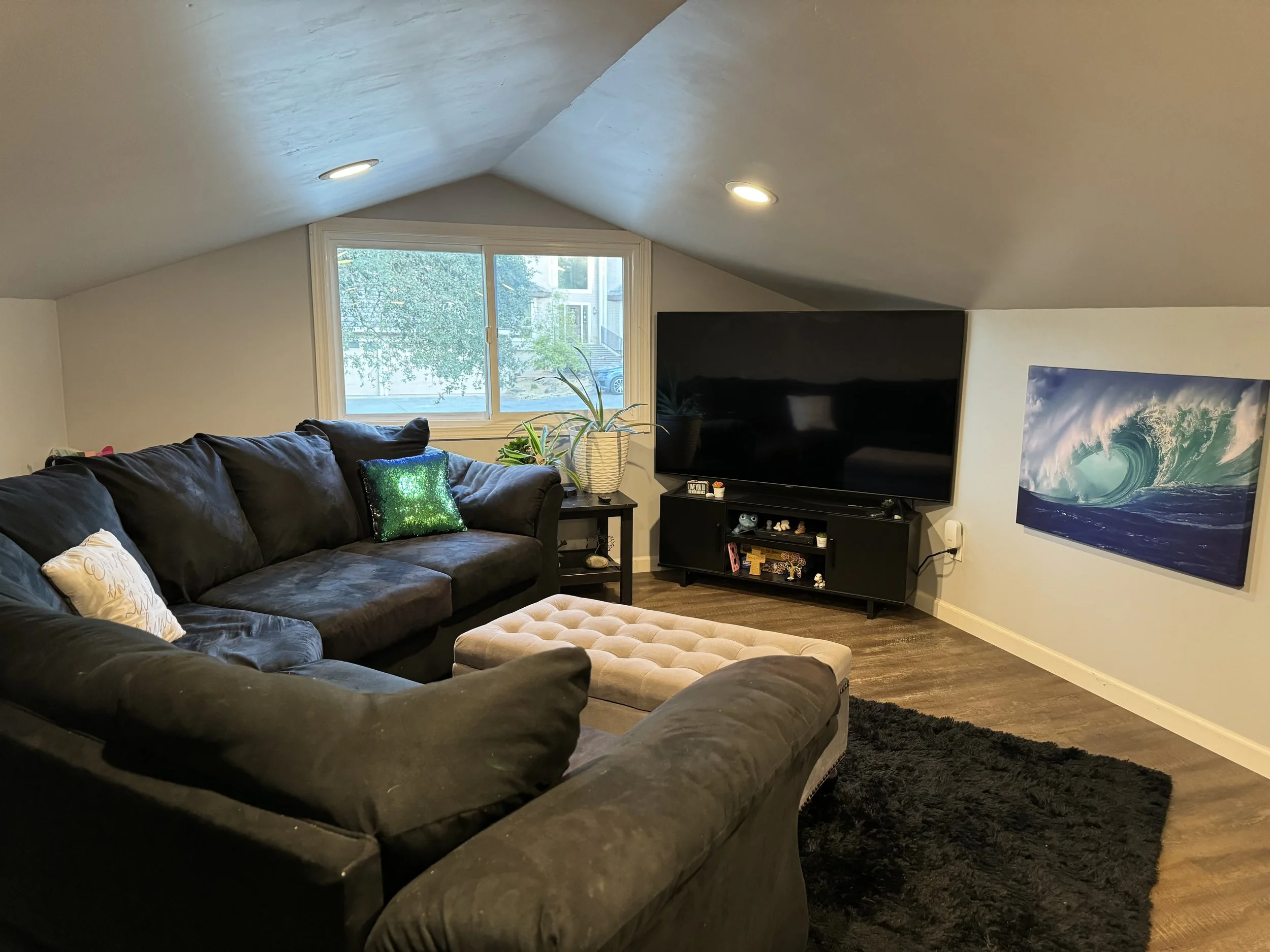 Living room with black sofa, beige ottoman, wall-mounted flat-screen TV, ocean wave artwork, window with outside view, and plants