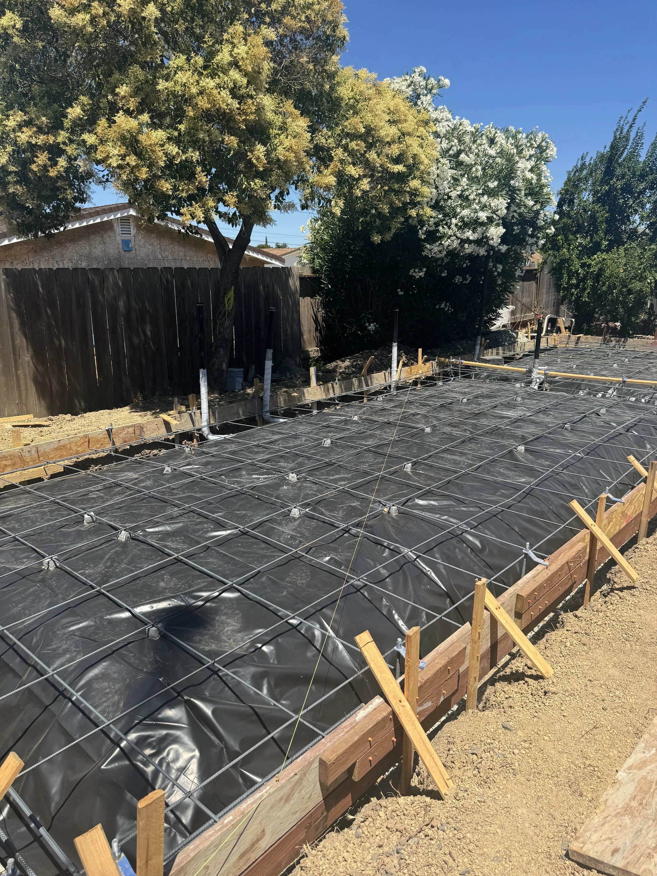 Construction site with rebar and black plastic sheeting in a foundation area, surrounded by wooden forms and sand, with two trees and a wooden fence in the background under a clear blue sky.