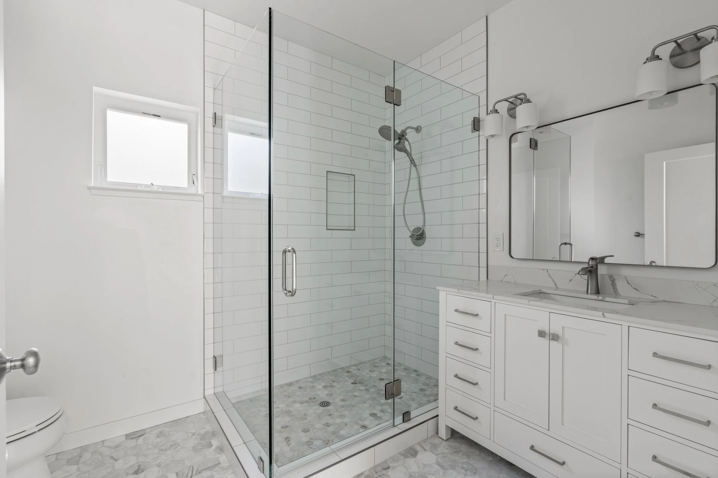 Modern white bathroom with glass shower enclosure, white cabinetry, marble countertop, and large mirror.