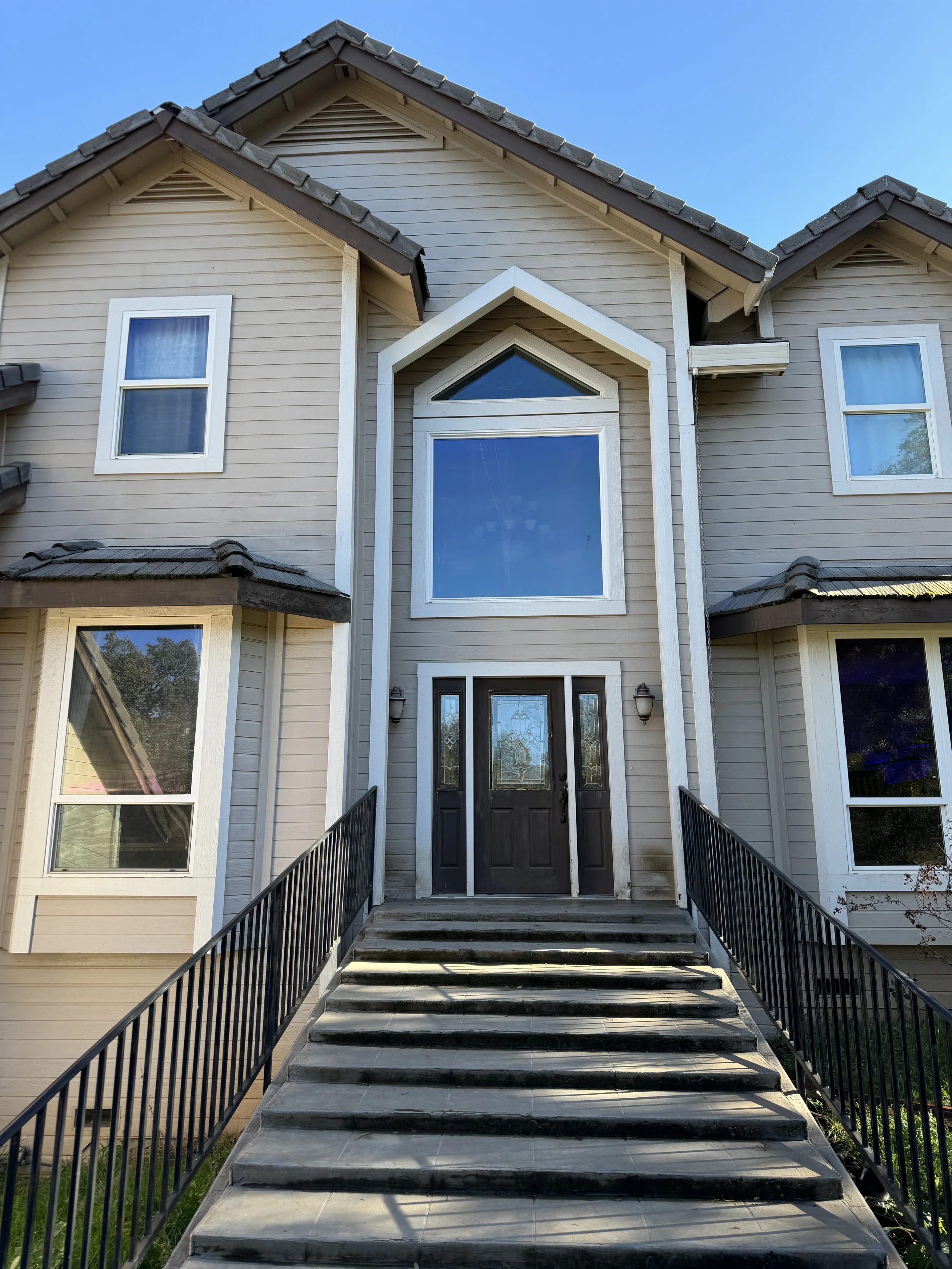 Front view of a modern two-story house with beige siding, black front door with glass, six steps leading to the entrance, and black railings on both sides. Windows with white frames are visible on the facade. The sky is clear and blue.