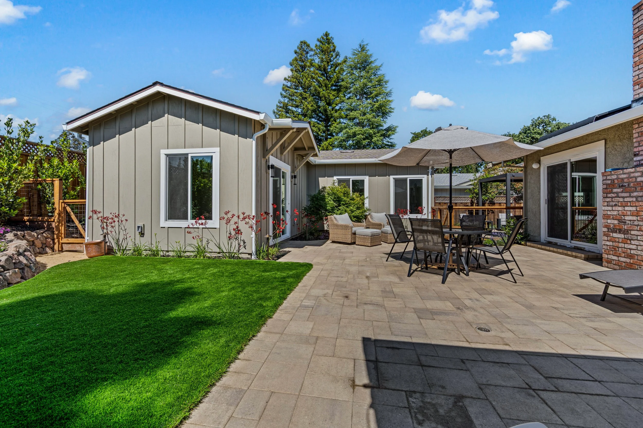 A backyard patio with outdoor furniture, a green lawn, and a wooden fence under a blue sky with a few clouds.