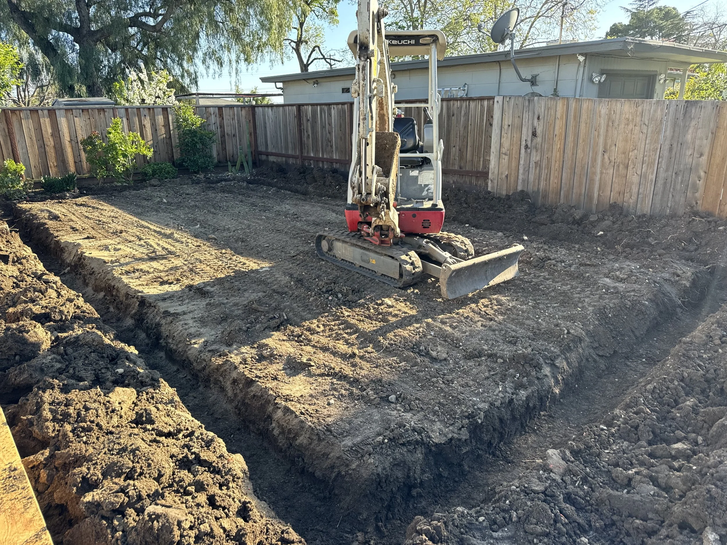 A small construction excavator working in a backyard, digging a trench for landscaping or utility installation, with a wooden fence, trees, and a house in the background.
