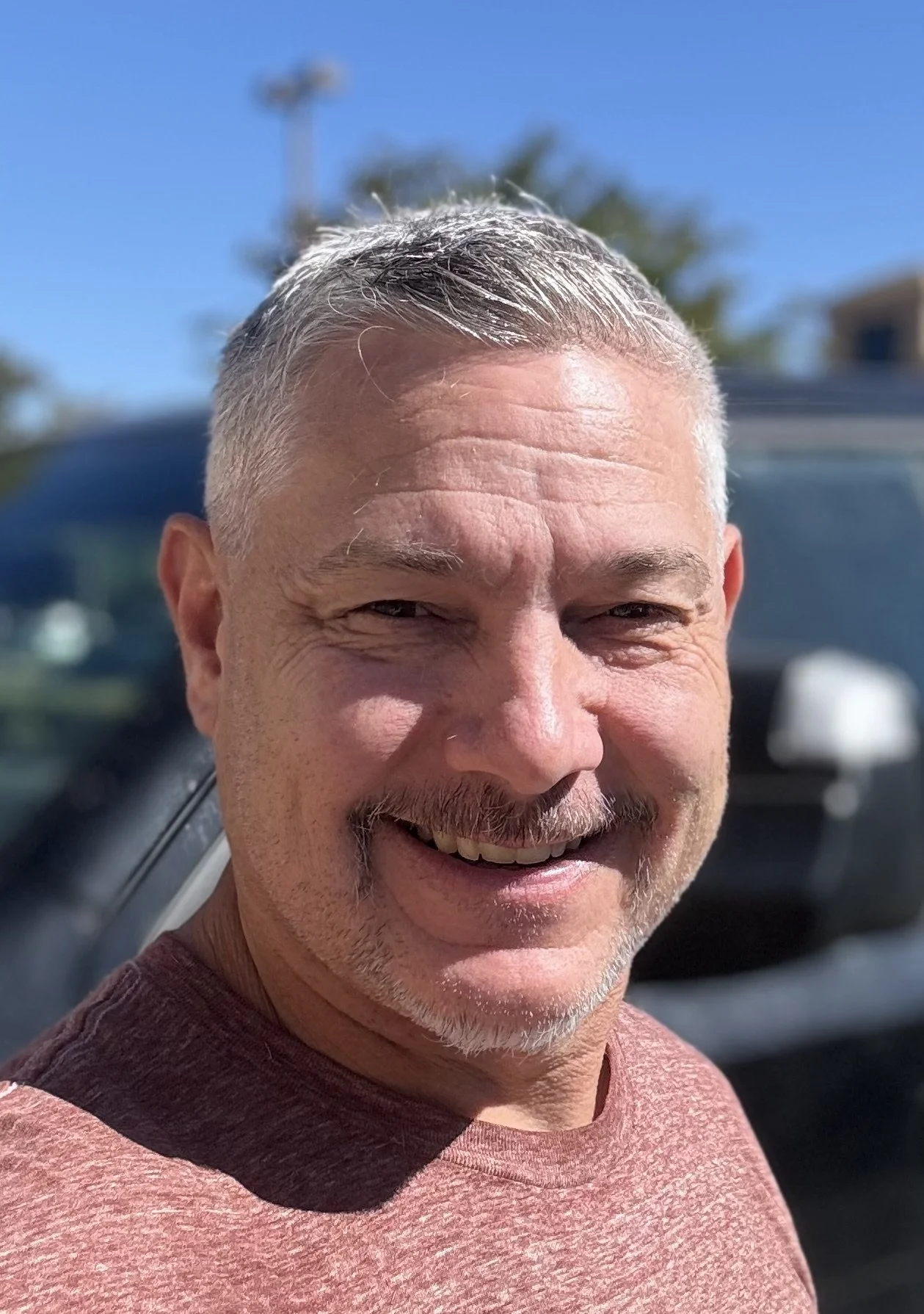 Close-up of a smiling man with short, gray hair and a beard, wearing a maroon shirt, outdoors on a sunny day.