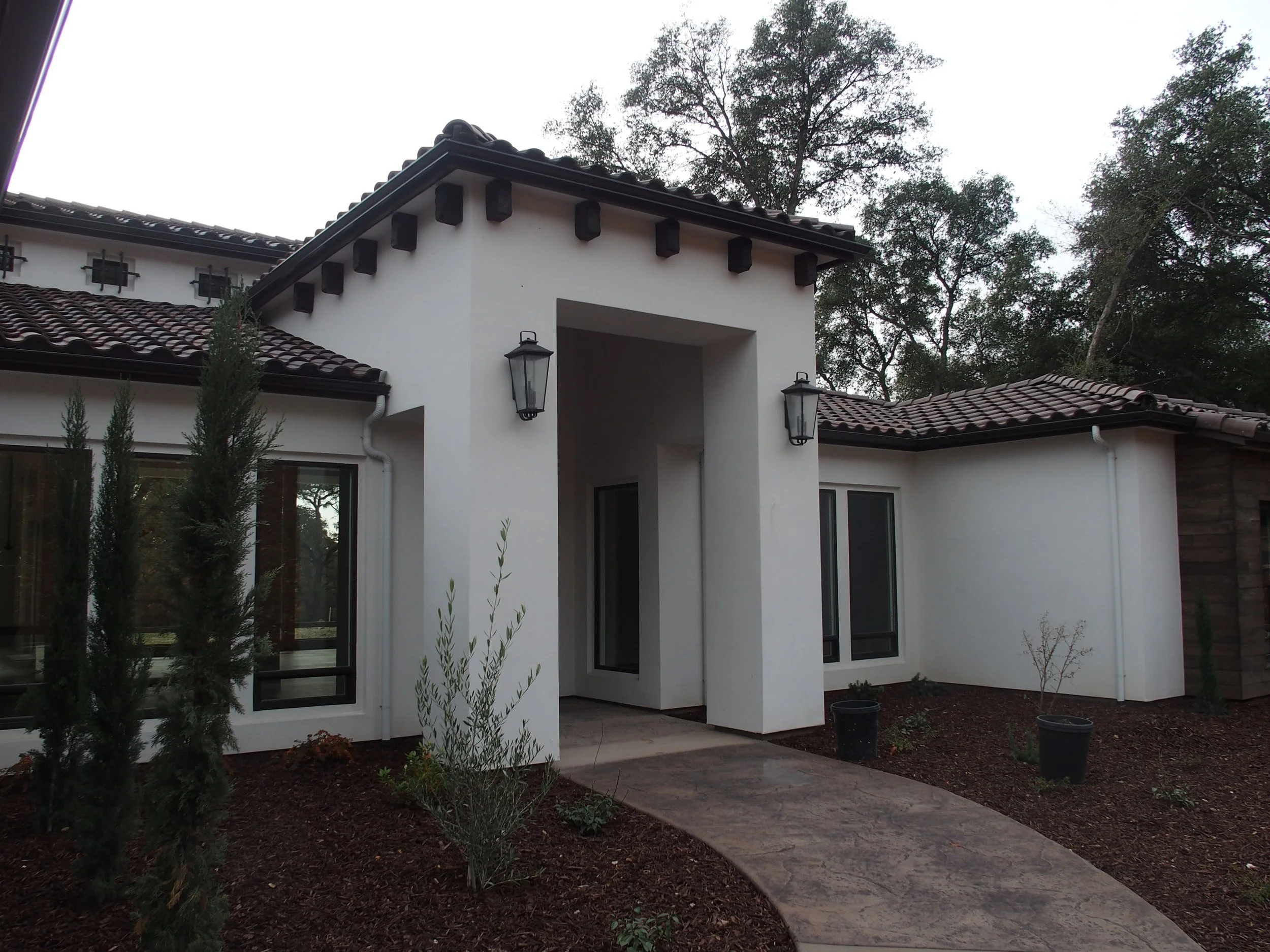 Front view of a modern house with white stucco walls, dark brown tile roof, and black lantern-style exterior lights. Small landscaped yard with shrubs and a curved walkway.