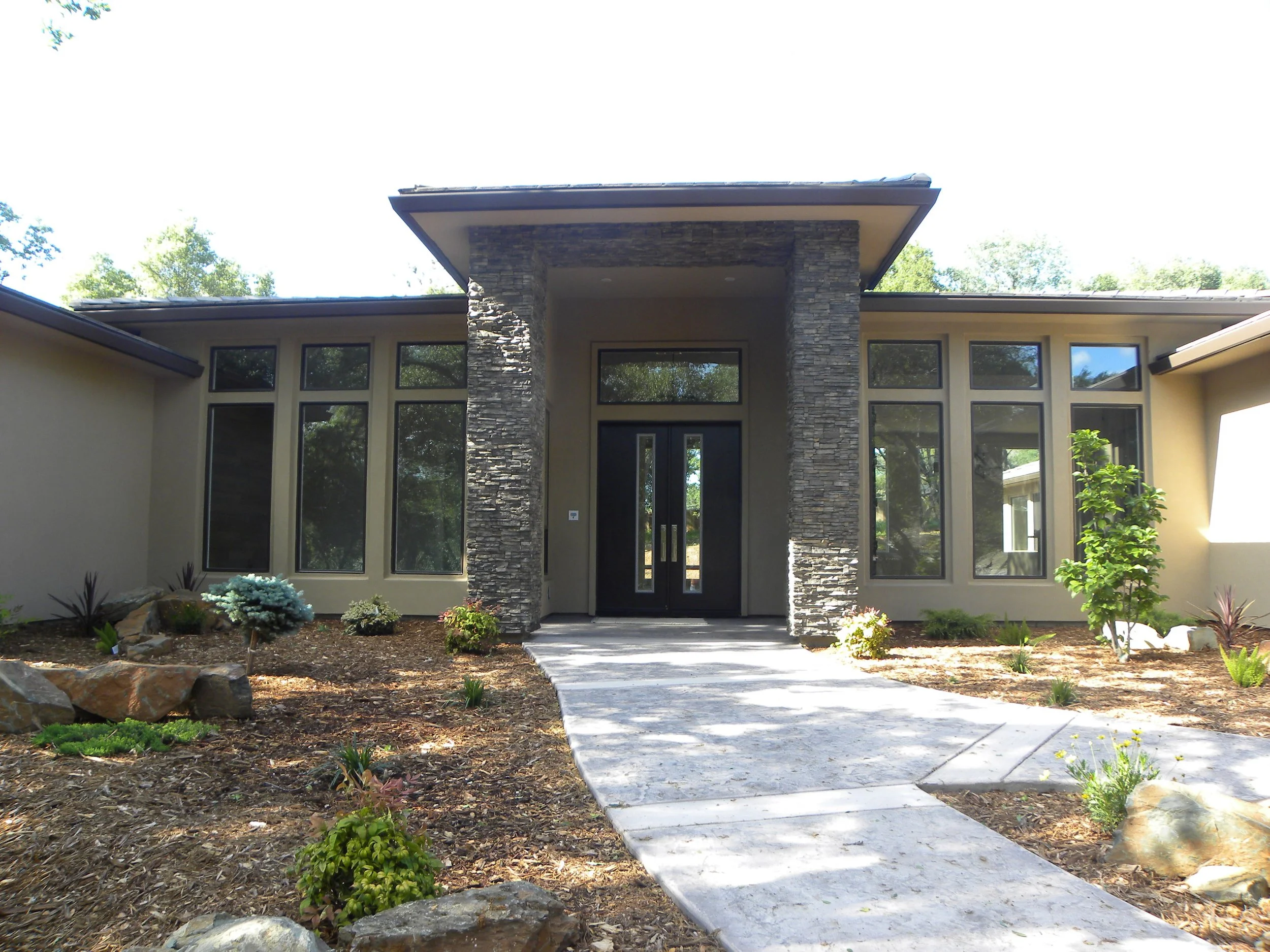 Front view of a modern house with a stone entryway, large windows, and a pathway leading to the door, surrounded by landscaping and trees.
