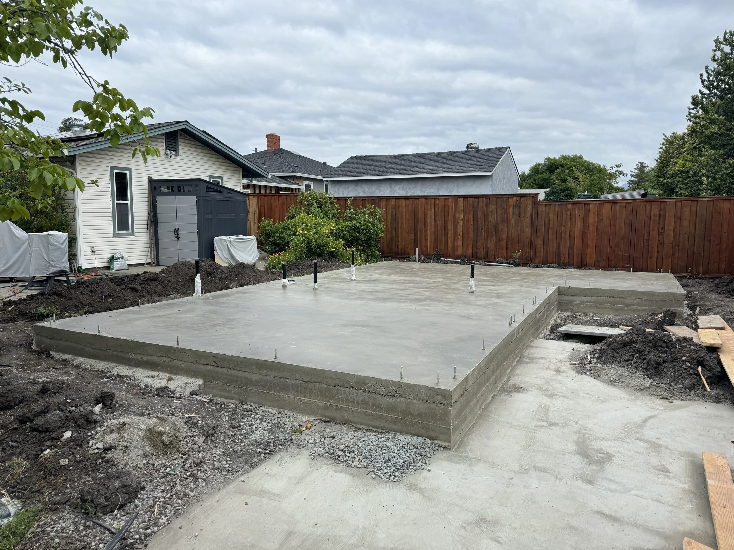 Freshly poured concrete slab in a backyard, surrounded by dirt, with a small staircase and partially visible wooden boards on the ground.