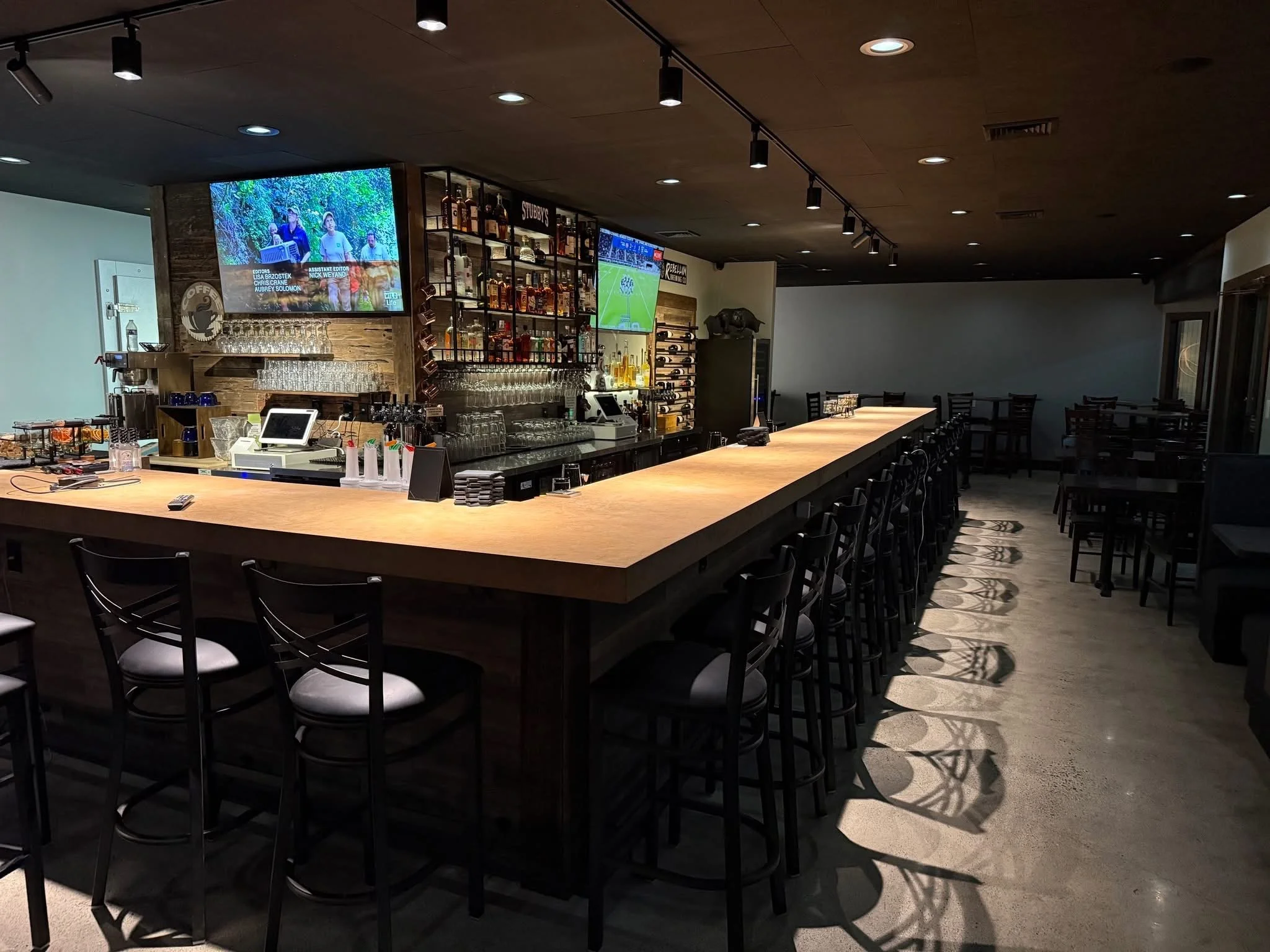 Empty bar with a long wooden counter and black chairs, shelves with bottles behind, TV screens displaying sports, and dim lighting.