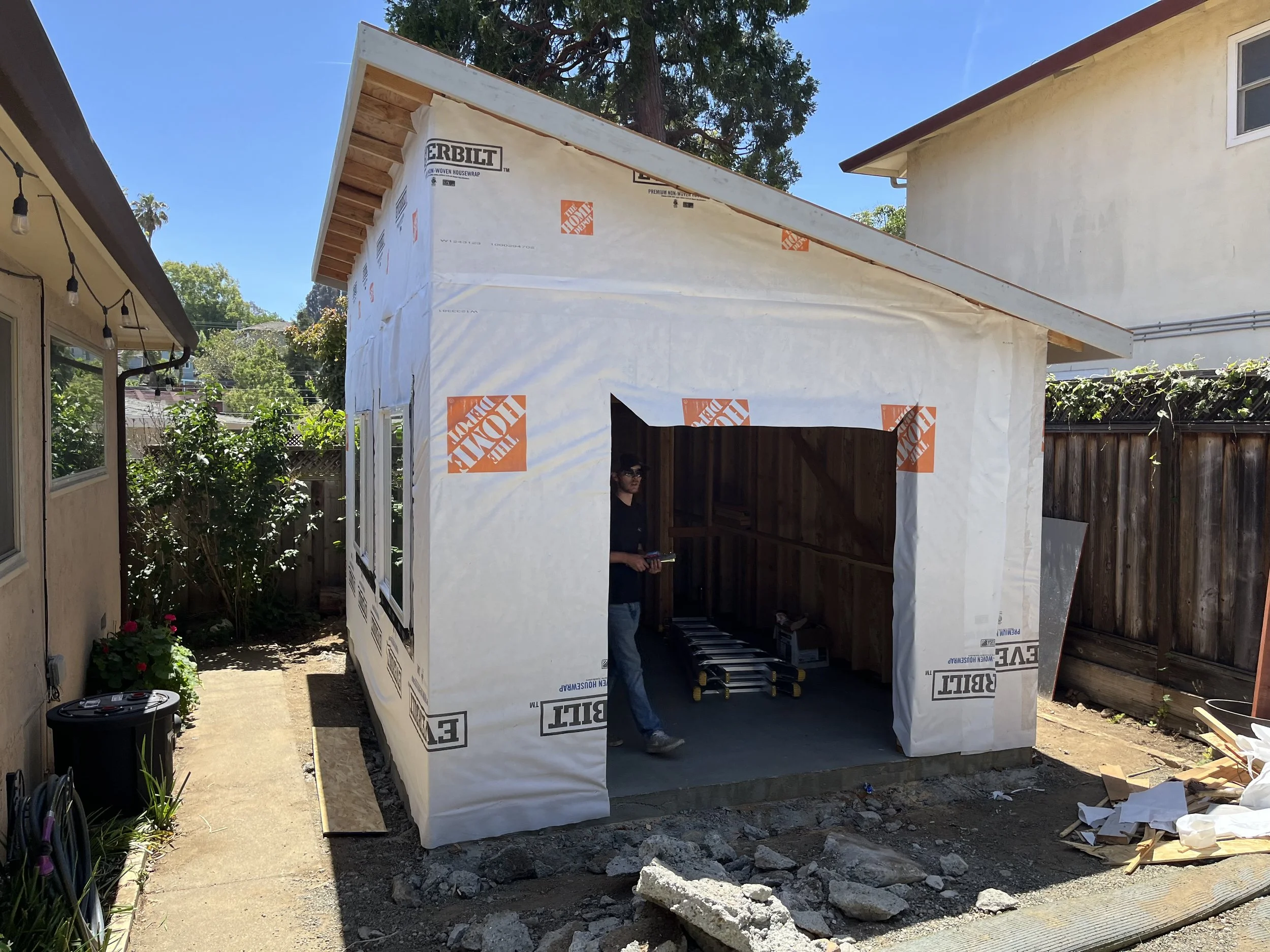 Small house under construction with wrap-around house wrap, a person standing inside, and construction materials on the ground.