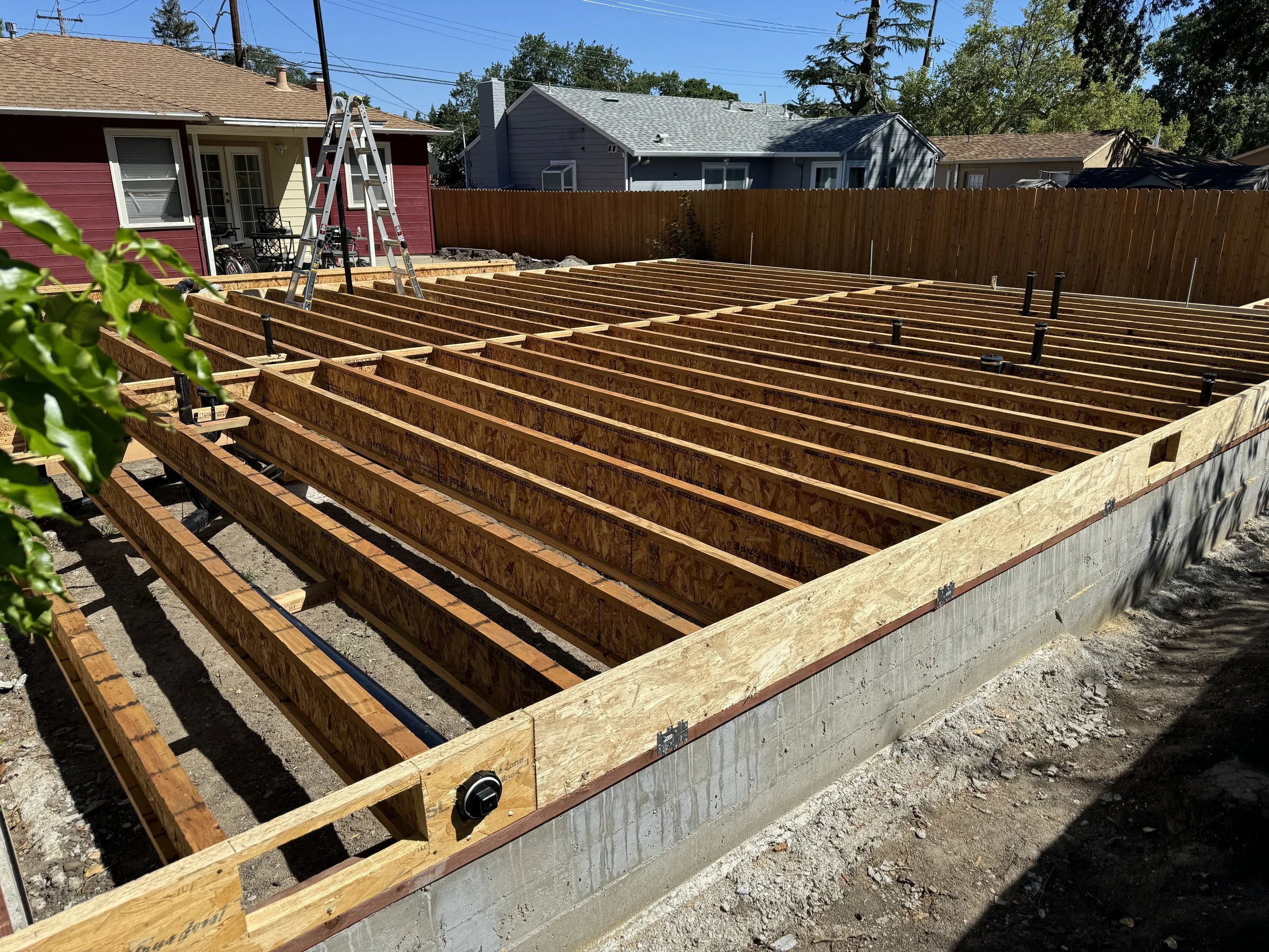 Construction site with wooden framing for a building, situated in a backyard with neighboring houses and a wooden fence.