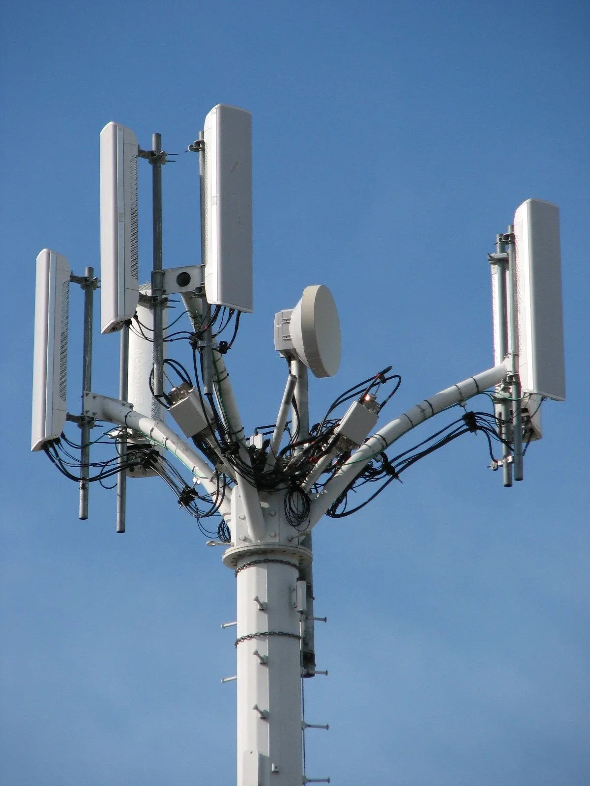 Cell tower with multiple antennas and cables against a blue sky.