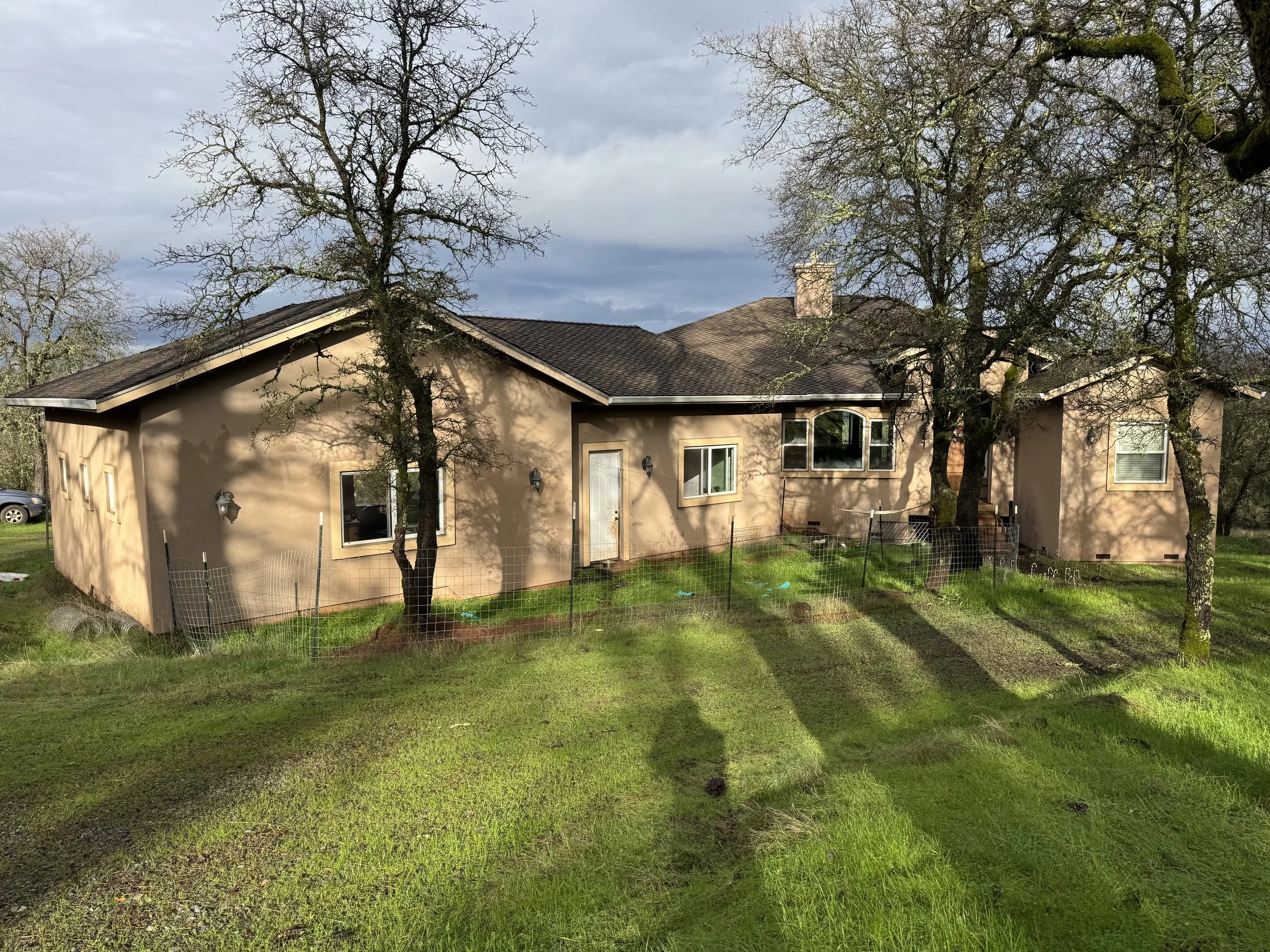 A single-story beige house with multiple windows and a white door, surrounded by a green lawn, with three leafless trees and a cloudy sky in the background.