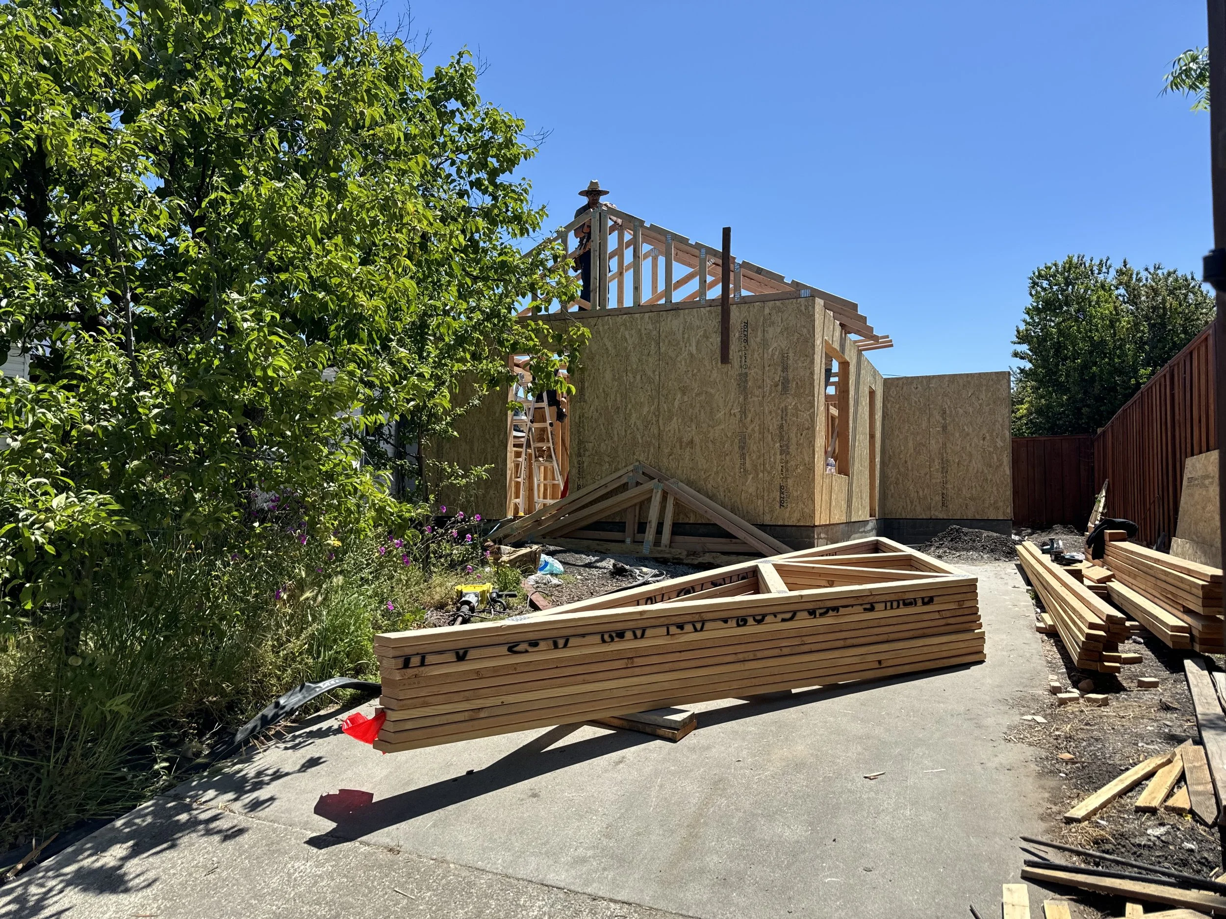 Construction site with partially built wooden house, stacks of lumber, and a worker on the roof under a clear blue sky.