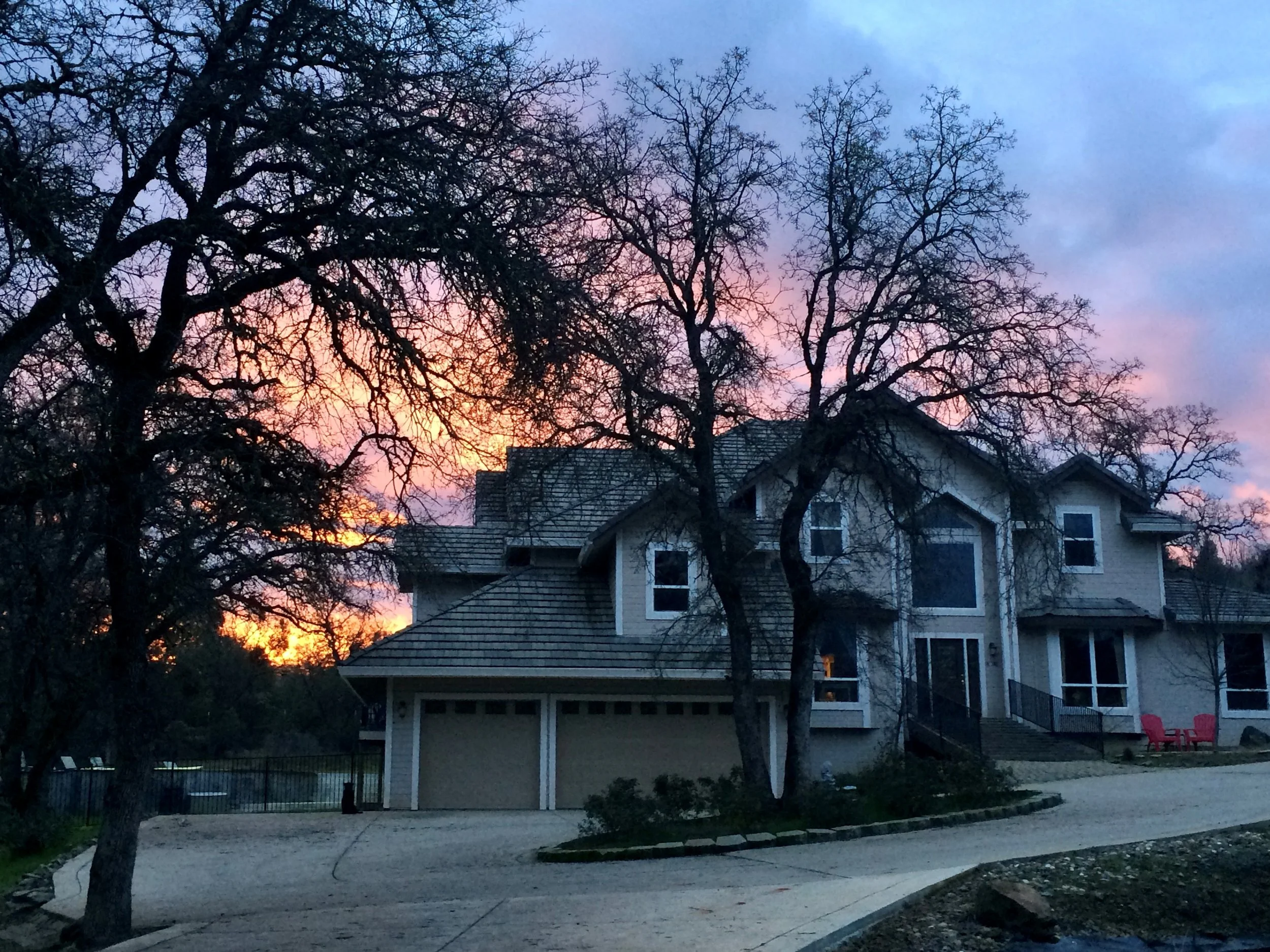 A two-story house with an attached garage, set against a colorful sunset with pink, purple, and orange sky. Bare trees frame the house, and there are two red chairs on the front porch.