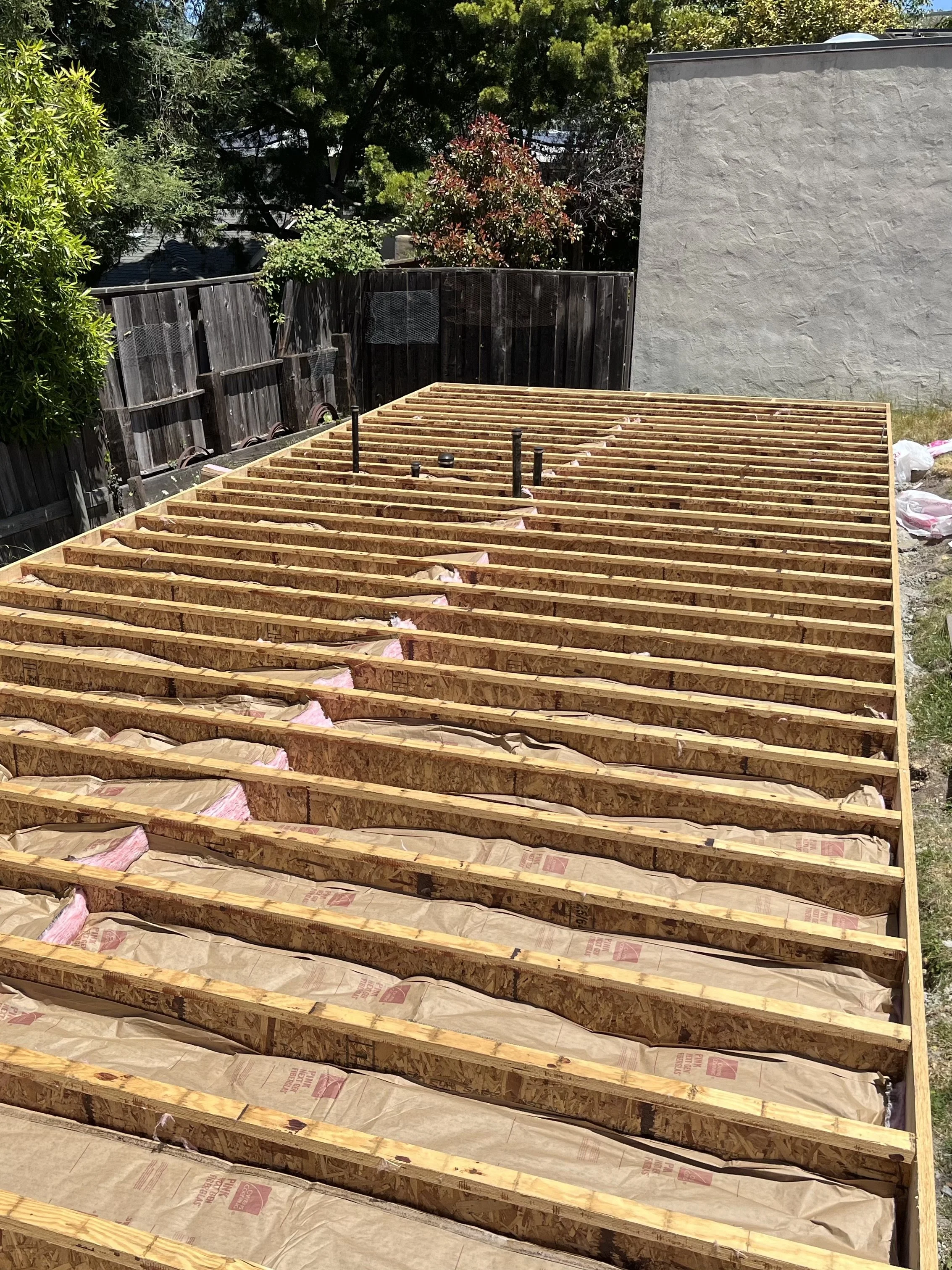 The photograph shows a construction site with wooden framing for a floor or deck, with insulation wrapped in paper laid between the joists. In the background, trees and a wooden fence are visible.