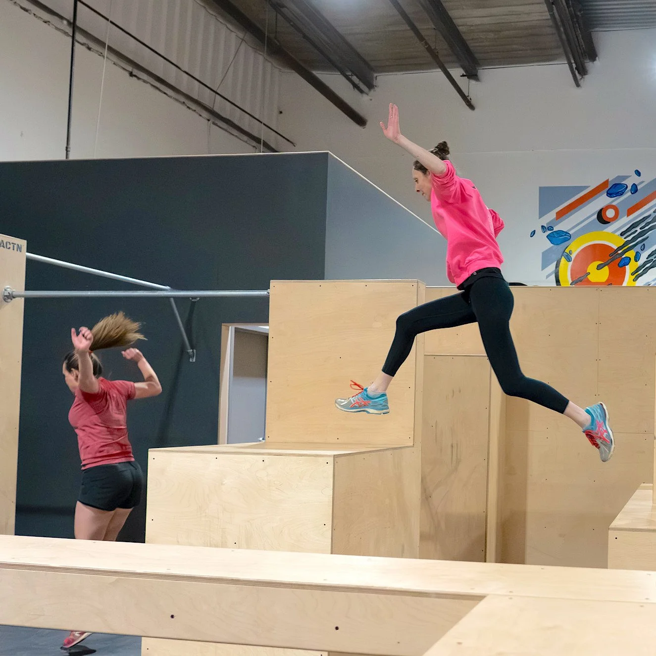 Deux femmes pratiquent le parkour dans un gymnase, sautant par-dessus des obstacles en bois, vêtues de vêtements de sport décontractés.
