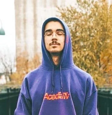 Un jeune homme avec des lunettes, portant un sweat à capuche violet avec le logo 'Accepttv', se tenant devant des arbres automnaux.