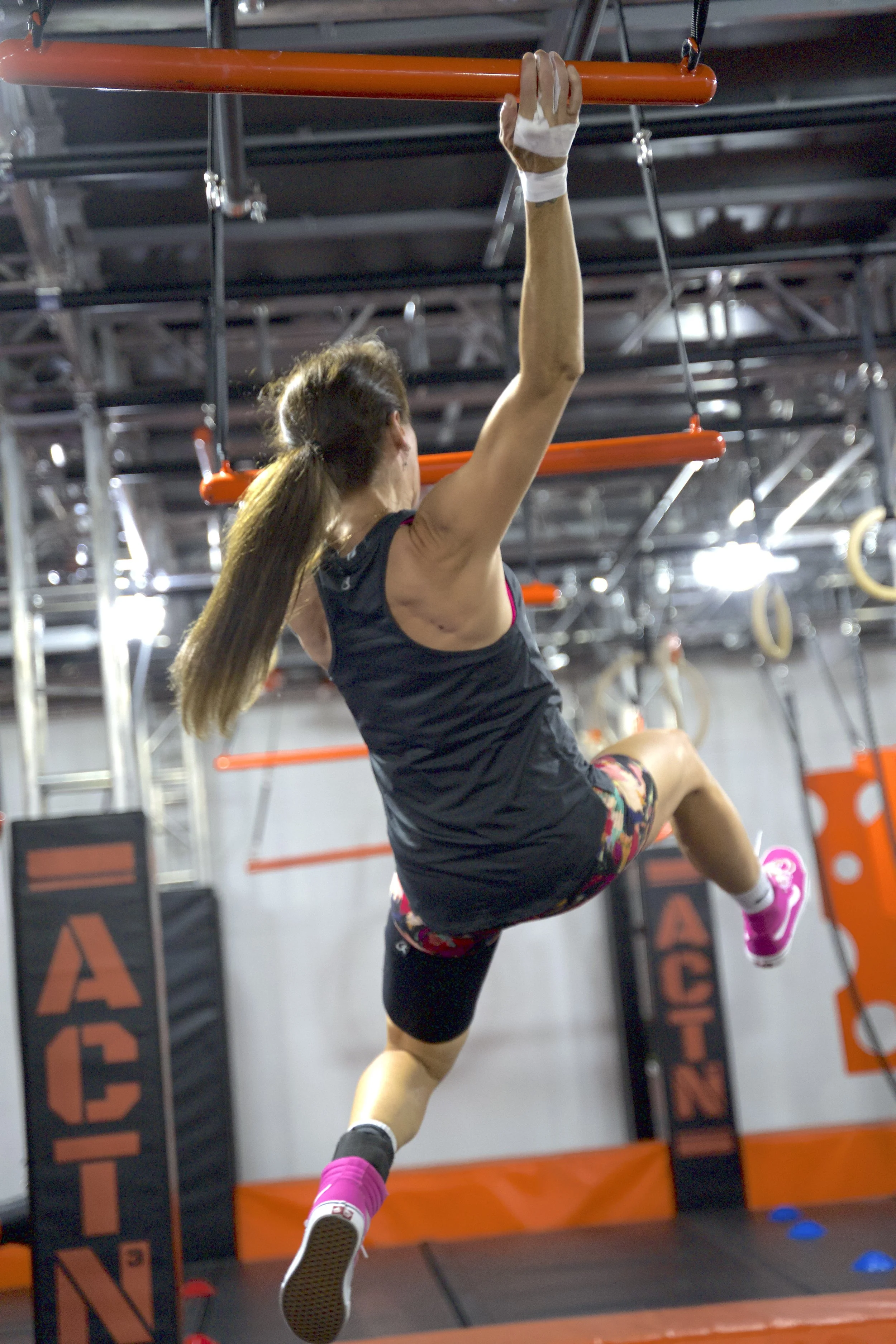 Une femme en vêtements de sport effectue un saut sur une plateforme dans une salle d'entraînement de Parkour ou Ninja Warrior, équipée de barres et cerceaux.