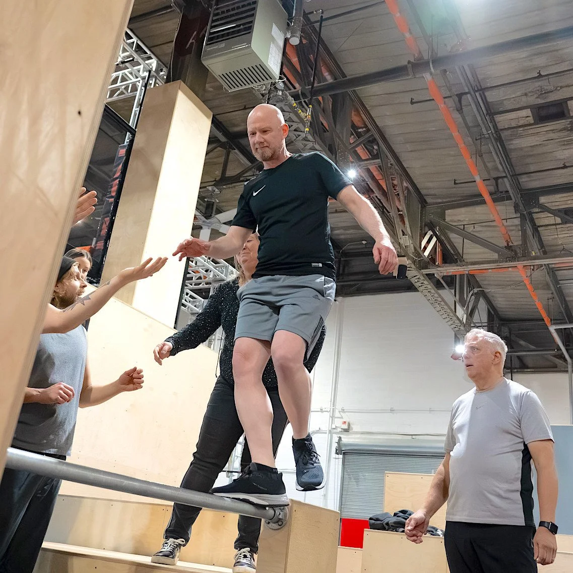 Un homme en tenue de sport noire traverse un obstacle en équilibre dans un centre d'entraînement intérieur. Plusieurs personnes l'observent et semblent prêtes à l'aider.