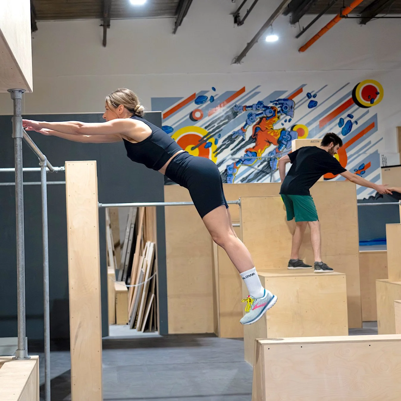 Deux personnes pratiquent le parkour dans un gymnase intérieur avec des structures en bois. Une femme en tenue de sport saute vers une barre métallique, tandis qu'un homme en arrière-plan grimpe sur des blocs en bois.