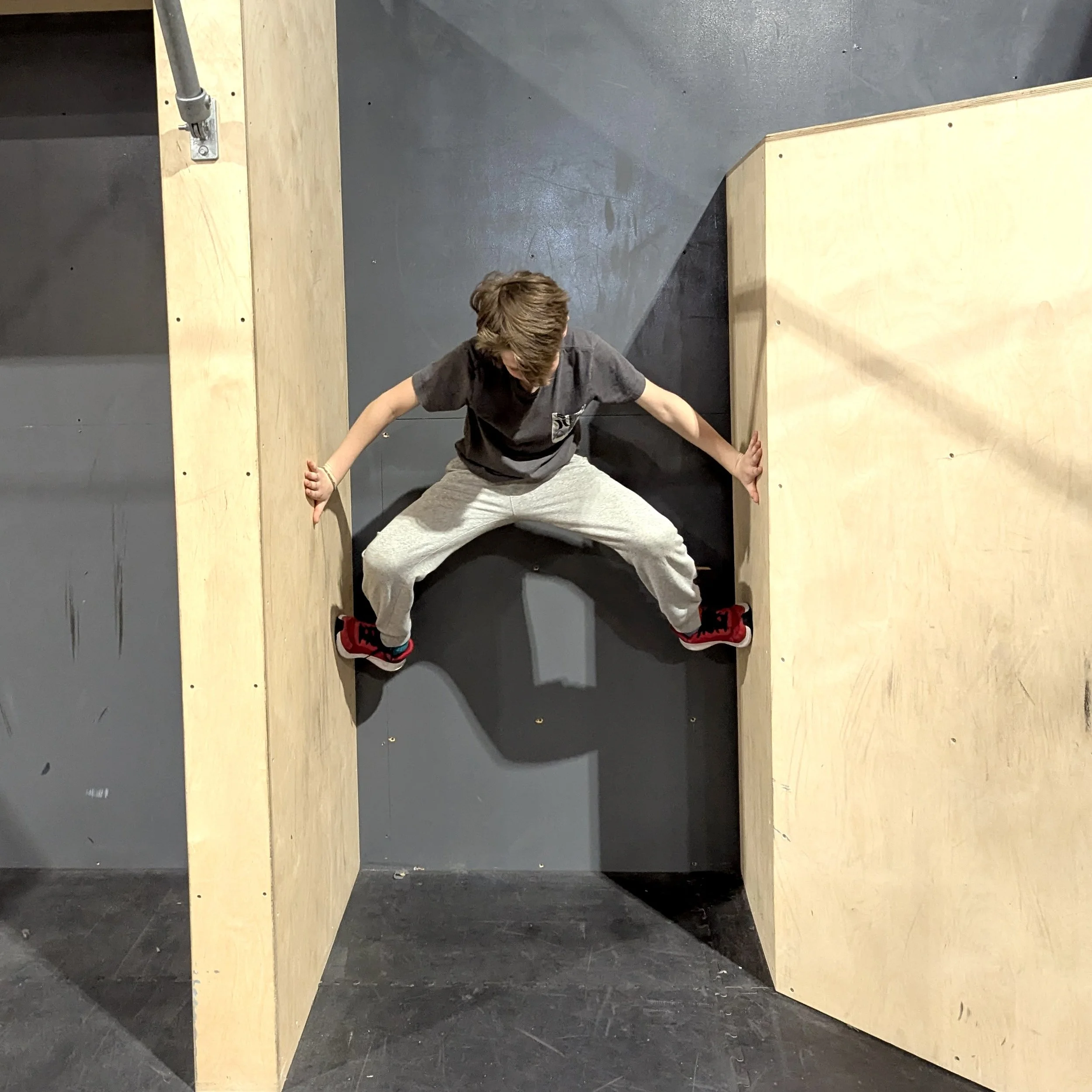Un garçon en tenue de sport tenant dans une posture de horizontale dans un espace intérieur avec des murs en bois et un sol en pierre.