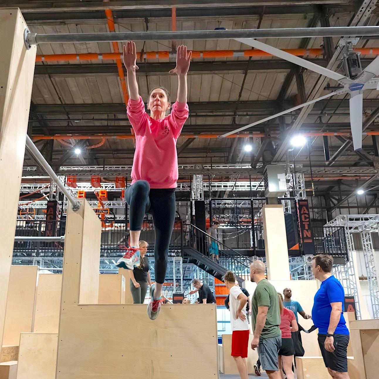 Une personne en sweat rose saute dans un environnement de gymnase intérieur avec des obstacles en bois. D'autres personnes se tiennent autour, regardant l'activité. L'espace est éclairé et équipé de structures métalliques pour l'entraînement physique
