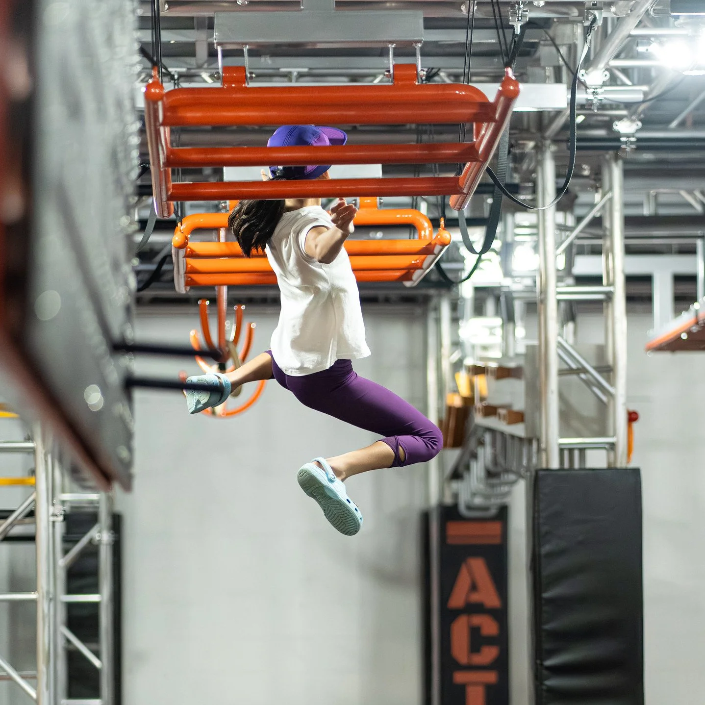 Une femme en train de faire du saut dans une salle d'escalade ou de parkour, suspendue sous un obstacle orange au plafond.