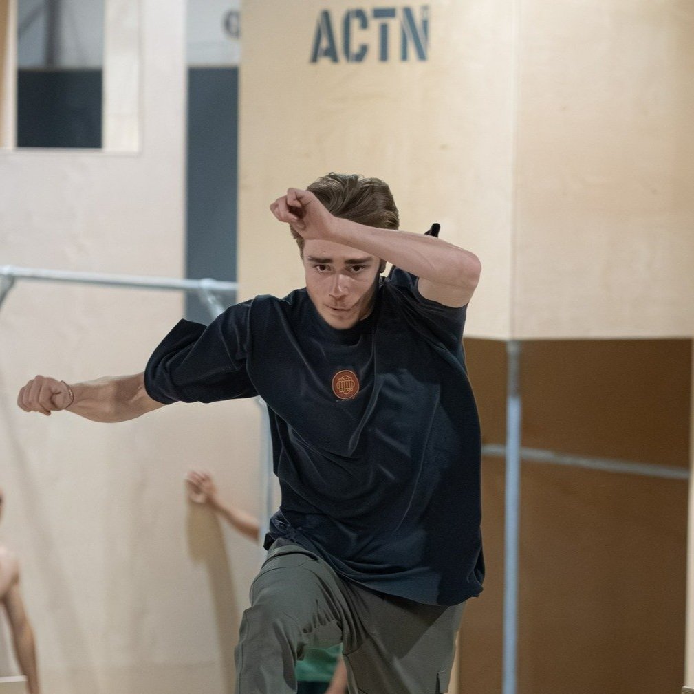 Un homme fait du parkour ou de la course d'obstacles en intérieur, portant un t-shirt noir et des pantalons gris, avec un mur marqué 'ACTN' en arrière-plan.