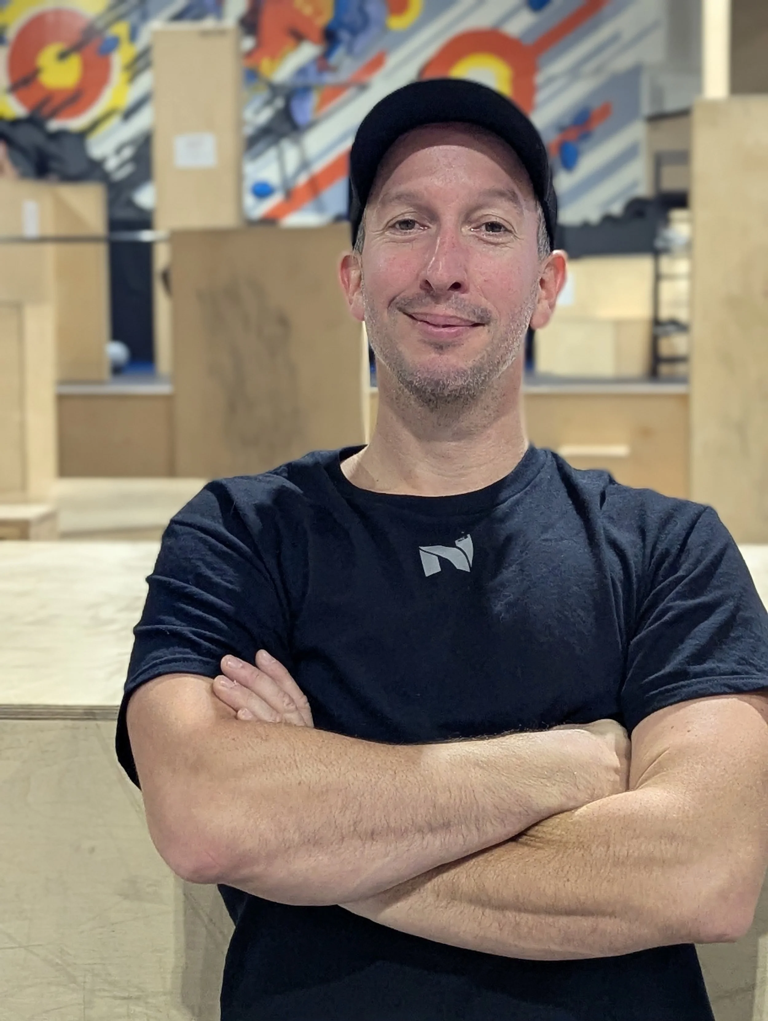 Homme souriant portant un t-shirt noir et une casquette noire, bras croisés, en arrière-plan un mur avec de l'art graphique coloré.